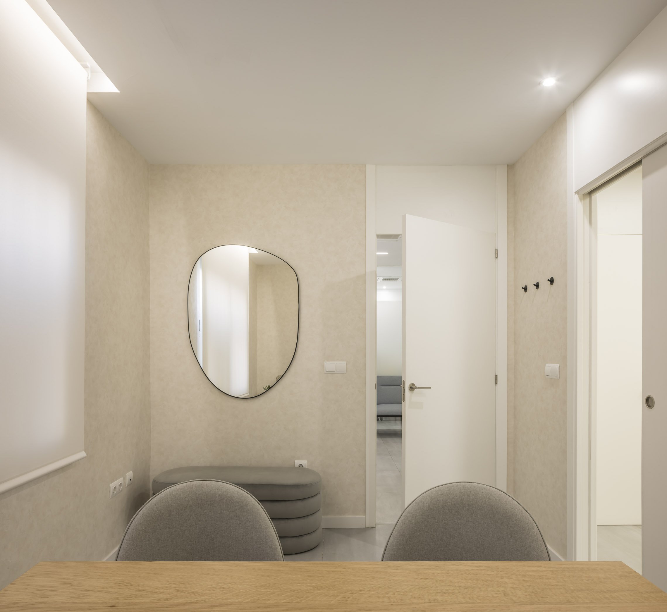 Vista interior de una sala moderna con una mesa de madera, sillas grises, un espejo de forma irregular y paredes blancas, con puertas blancas y luz suave.