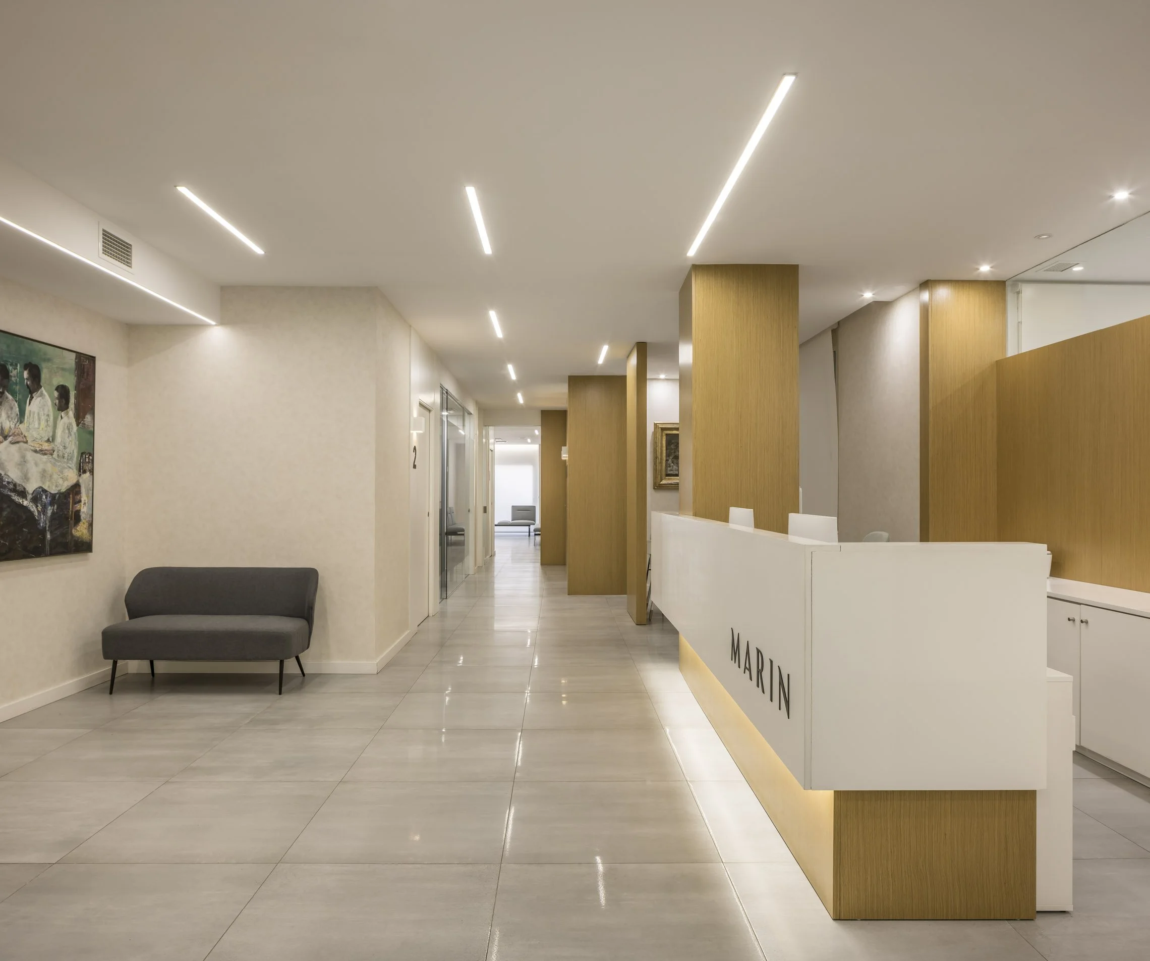 Recepción moderna con paredes blancas, divisiones de madera, cartel que dice 'MARIN' y área de espera con sillón gris y pintura en la pared.