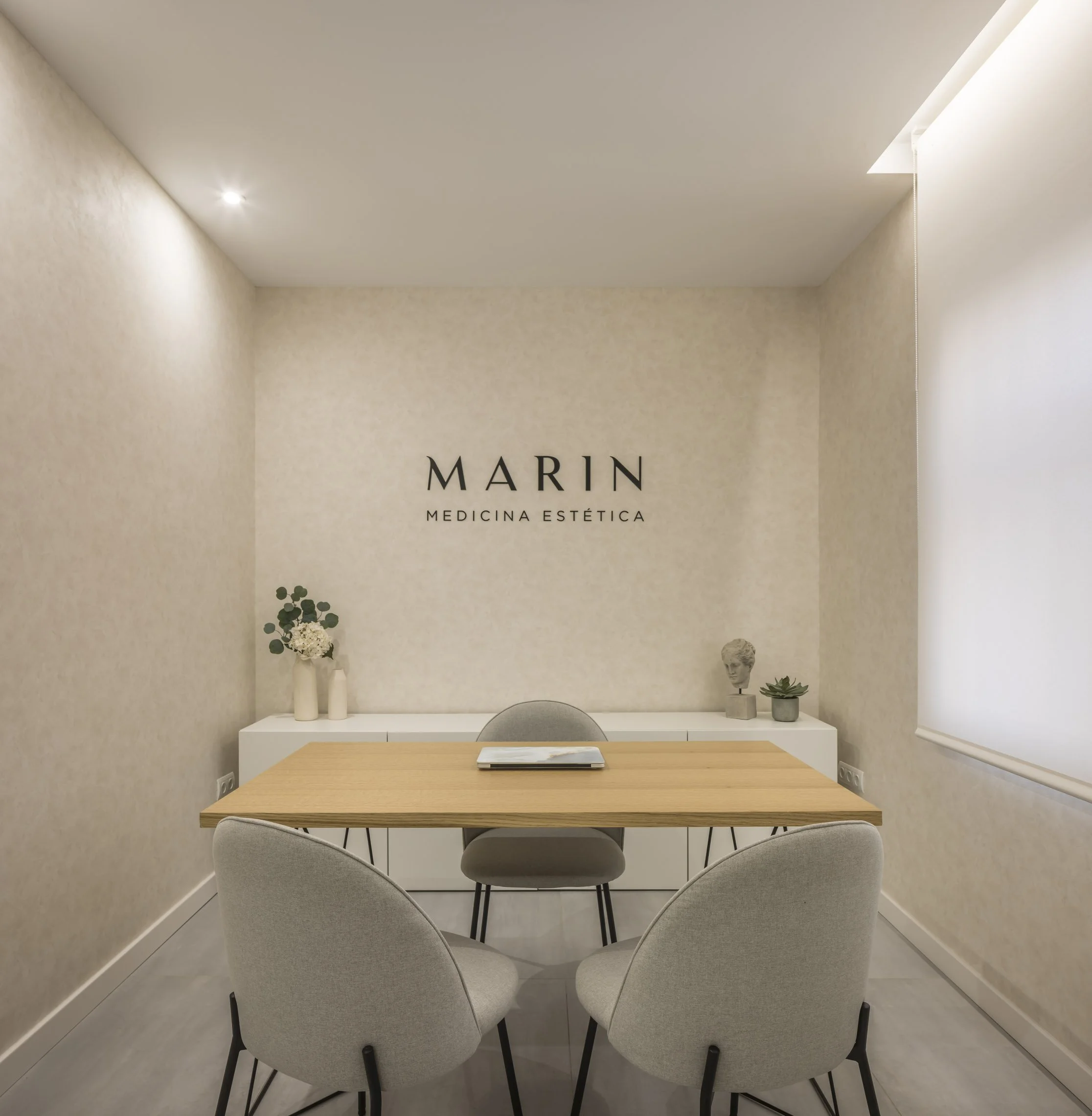 Sala de consultorios o sala de reunión con mesa de madera, seis sillas grises, paredes beige y decoraciones minimalistas con plantas y busto en una repisa. La pared tiene un cartel que dice "Marin Medicina Estética".