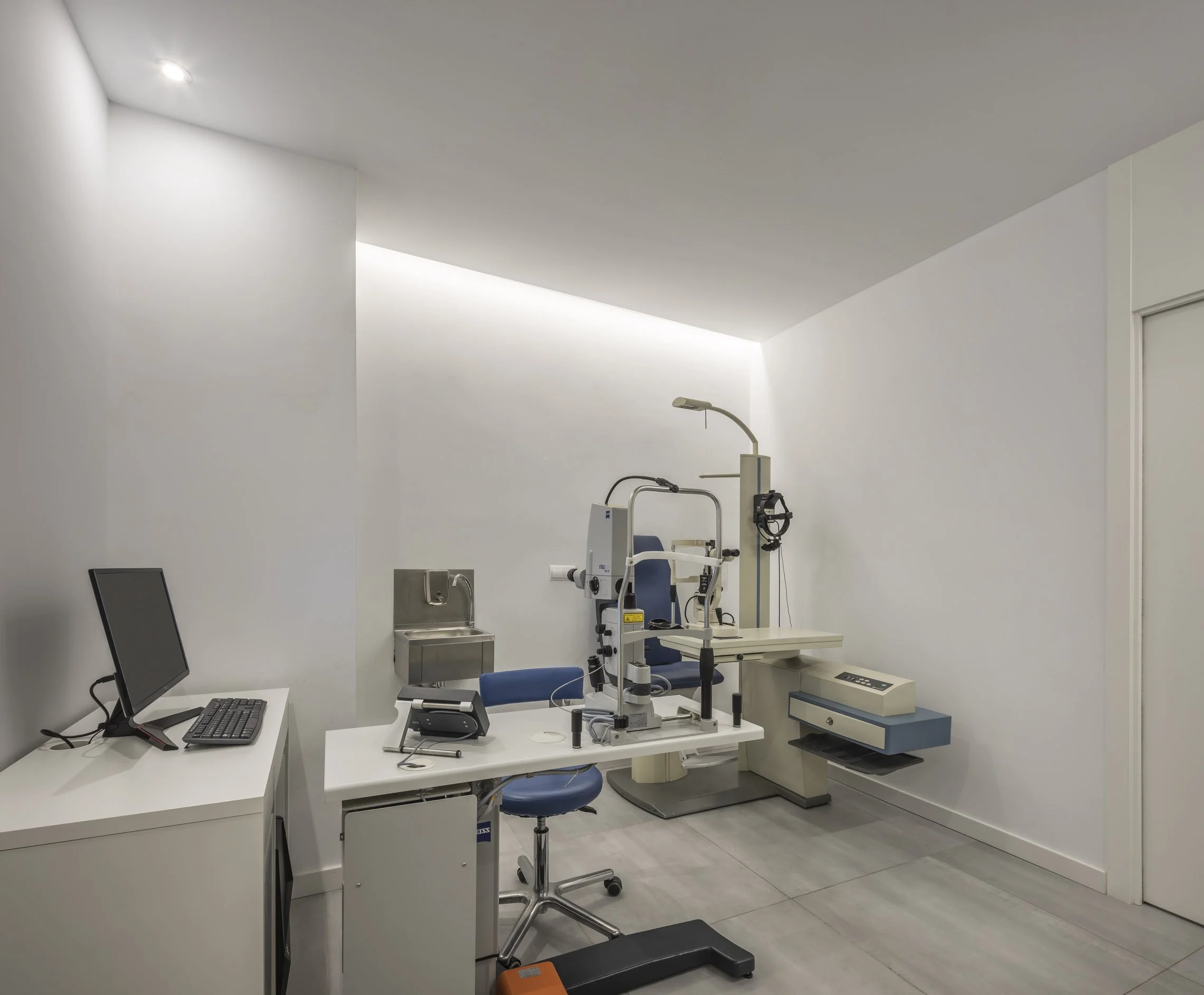 Sala de consulta médica con equipo de oftalmología, escritorio con computadora y sillas en un ambiente limpio y moderno.