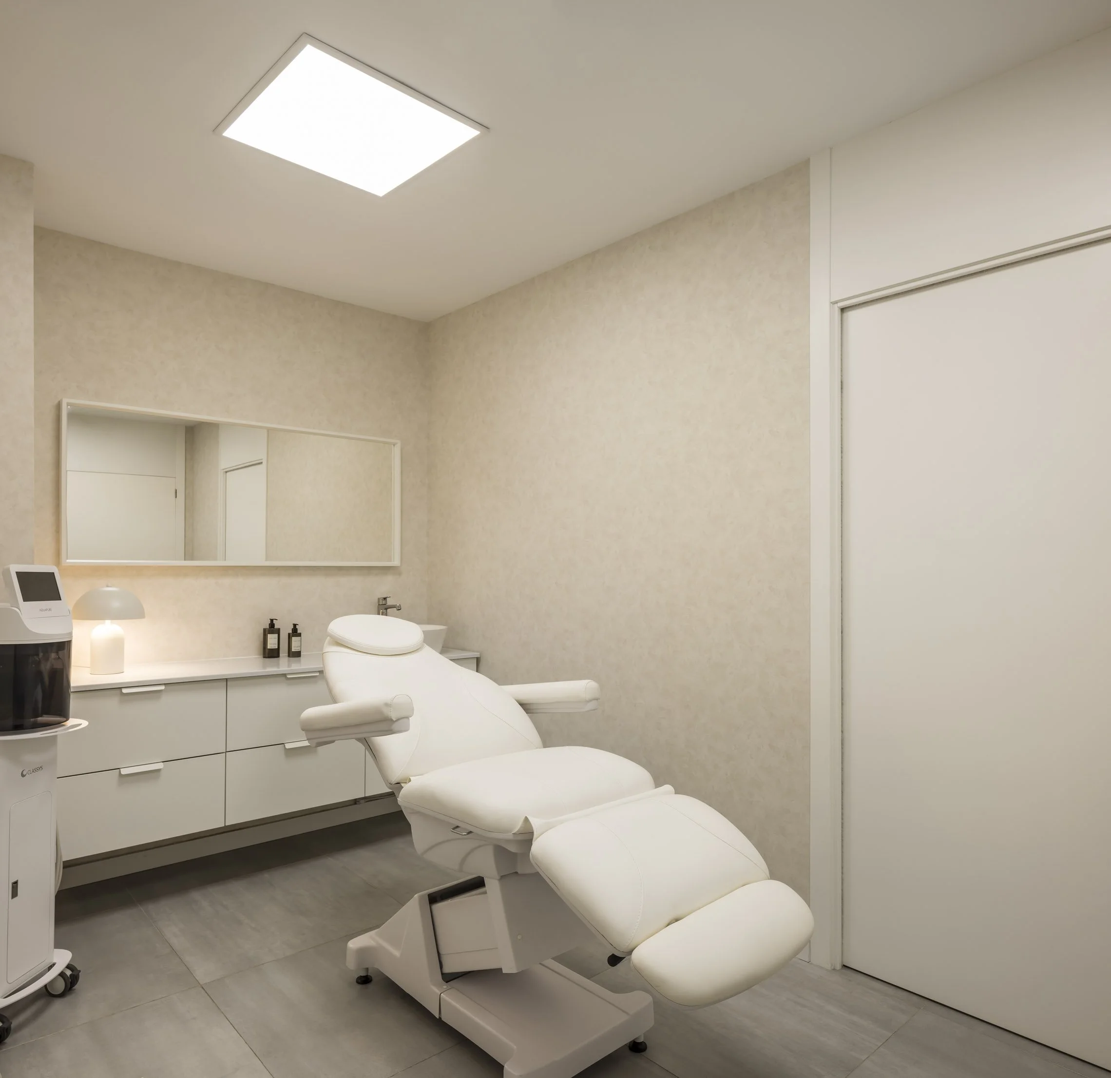 Sala de consulta en un consultorio médico con una cama, una lámpara de luz brillante en el techo, un espejo grande en la pared y un área con dispensadores y un robot de atención médica.