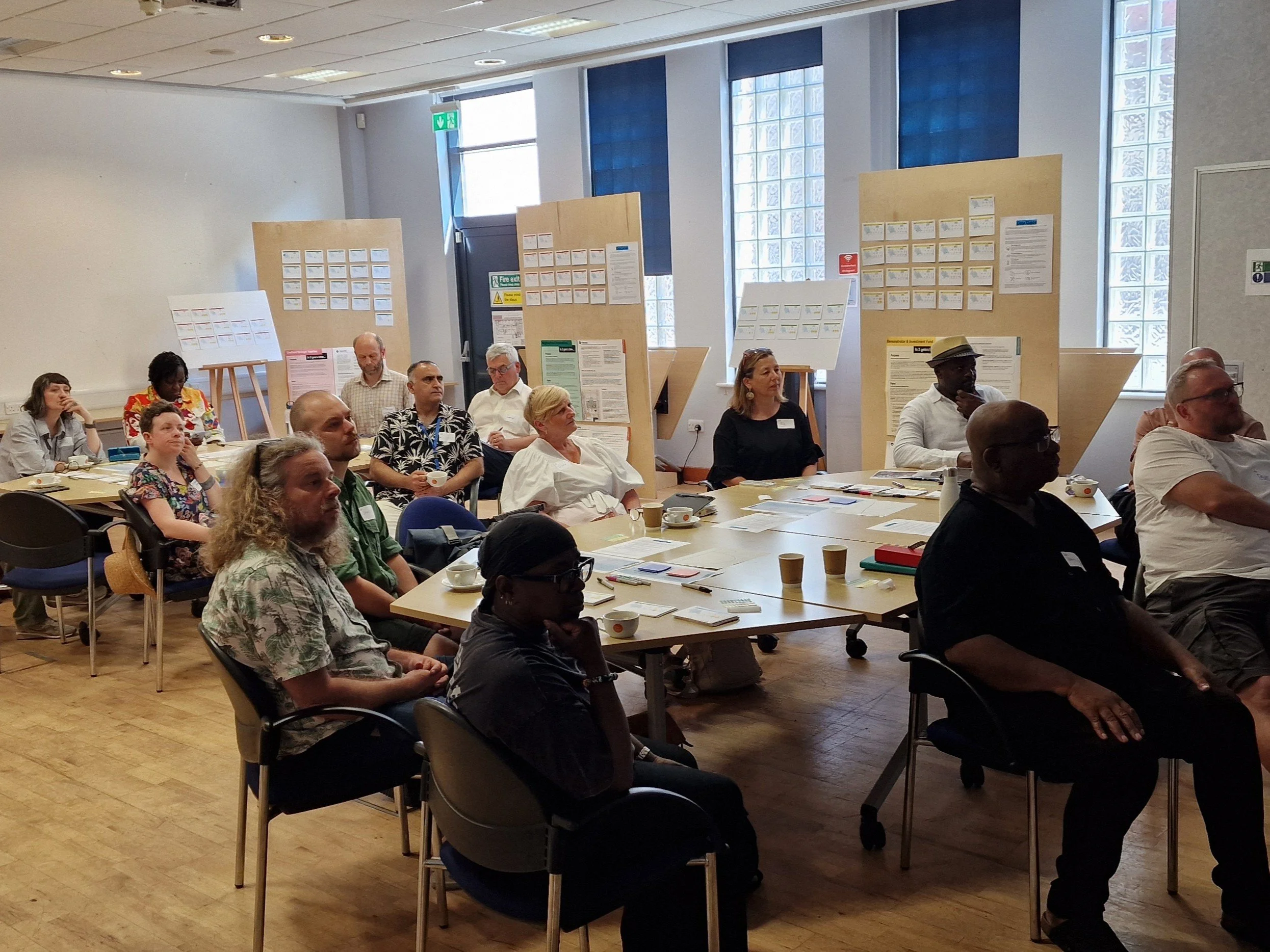 A photo of about 15 people sat around tables in a room, listening to a speaker out of shot