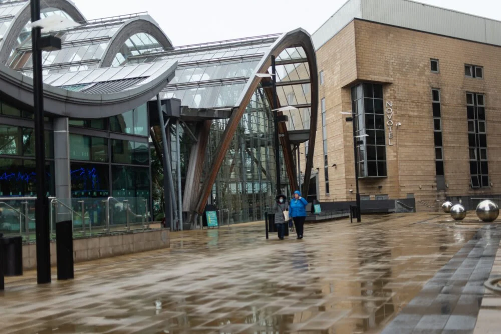 A photo taken on a rainy day, of a woman walking alone through an urban setting with buildings on either side of her