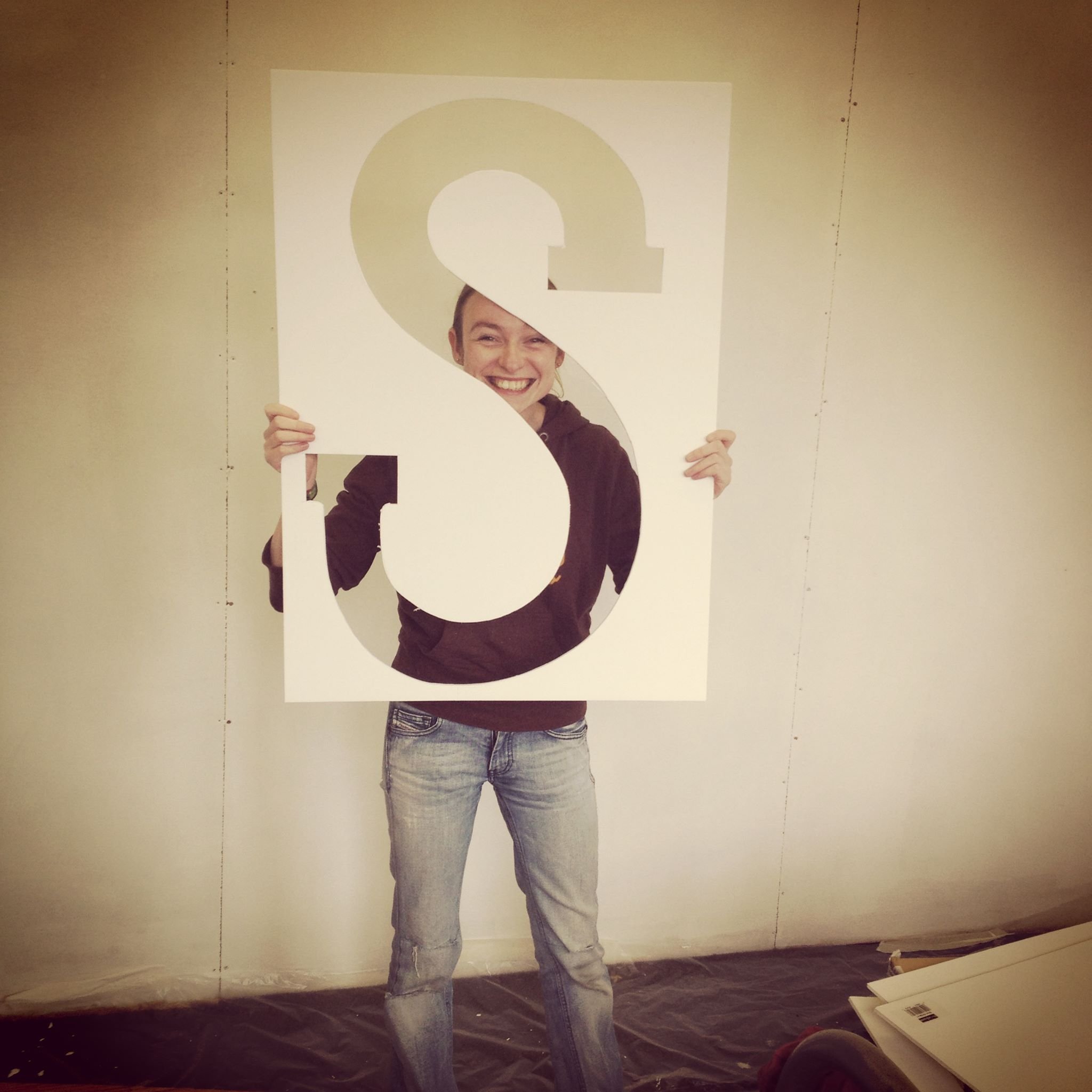 Sara holds up a large piece of paper with an 'S' cut out of it. She is smiling.