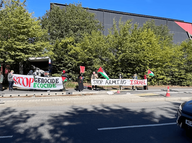 A photo of a group of around a dozen activists staging a protest at the side of a road. Some are waving Palestinian flags and others are holding banners that say 'STOP GENOCIDE ECOCIDE' and 'STOP ARMING ISRAEL'