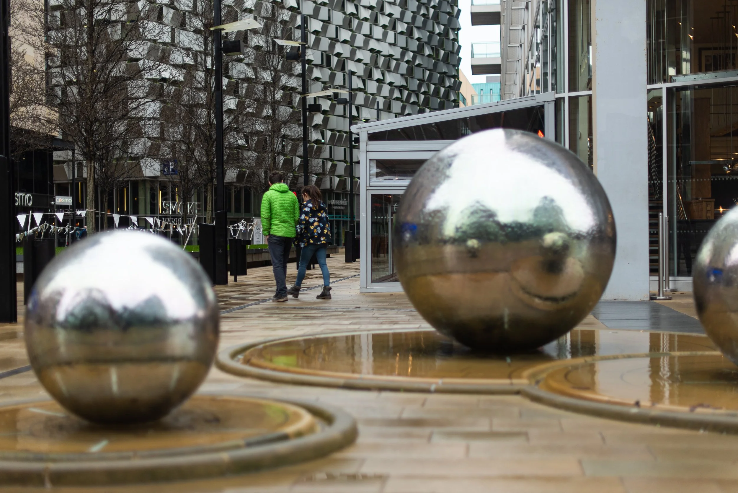 A photograph showing two water features in Sheffield city centre, which resemble large ball bearings. In the background you can see a number of modern buildings, and two people walking past.