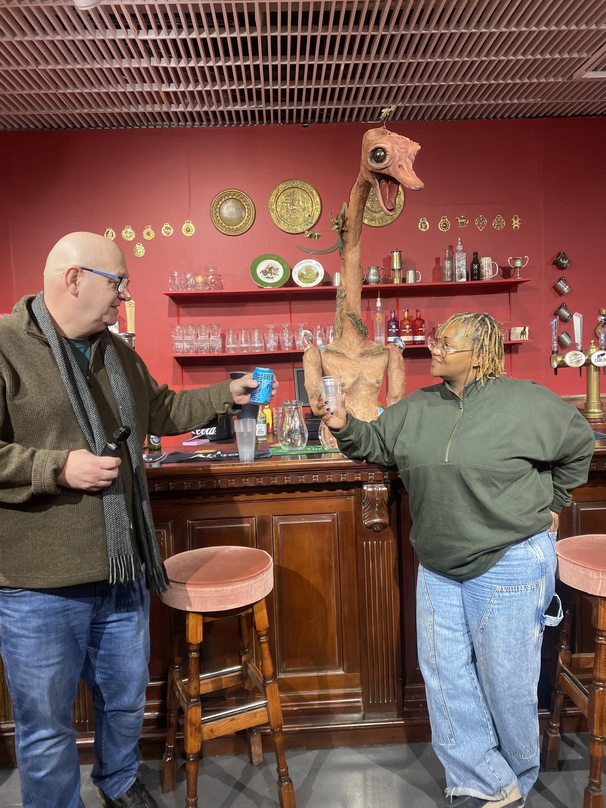 Two people stand at a bar, holding drinks. Behind the bar is a surreal sculpture showing a human torso with an ostrich's head.