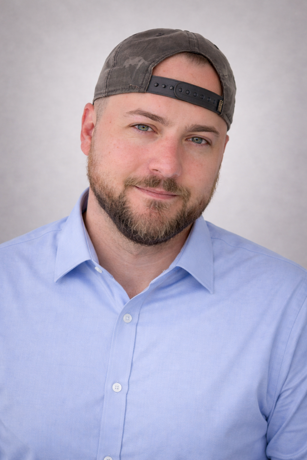 Portrait of a man wearing a blue button-up shirt and a backward baseball cap, with a short beard and light-colored eyes, smiling softly against a neutral background.