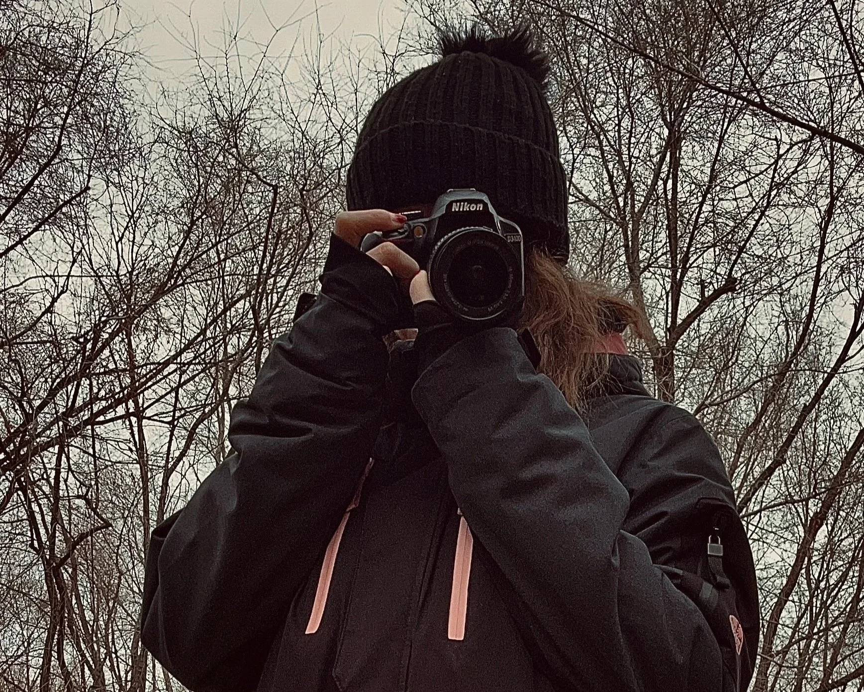 Persona con gorro negro y chaqueta negra sosteniendo una cámara Nikon, en un entorno de árboles sin hojas.