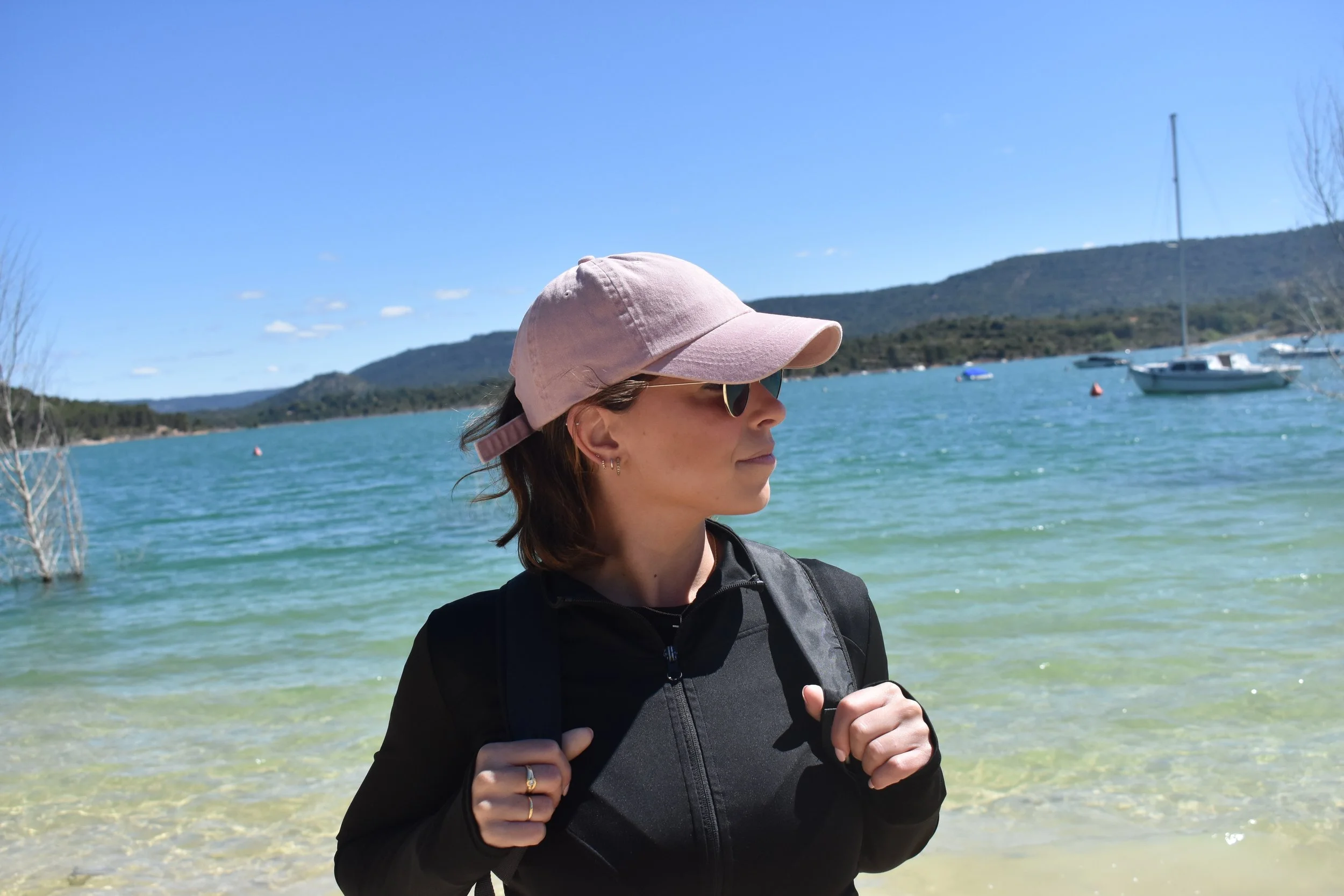 Mujer con sombrero rosa, gafas de sol y chaqueta negra, de perfil, en un entorno lacustre con barcos y montañas al fondo, durante el día con cielo despejado.