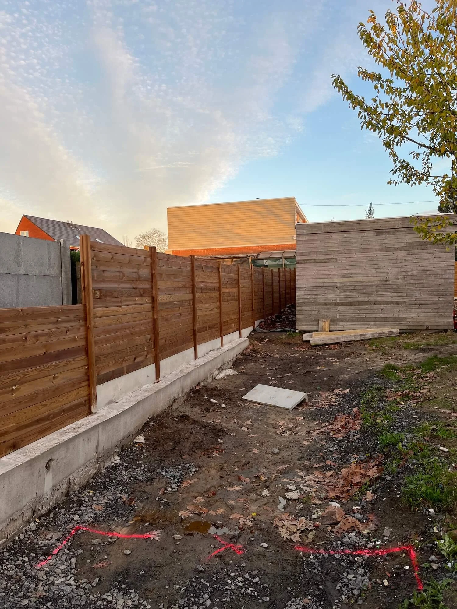 A backyard with a newly installed wooden fence, dirt ground with construction markers, and a partly cloudy sky.