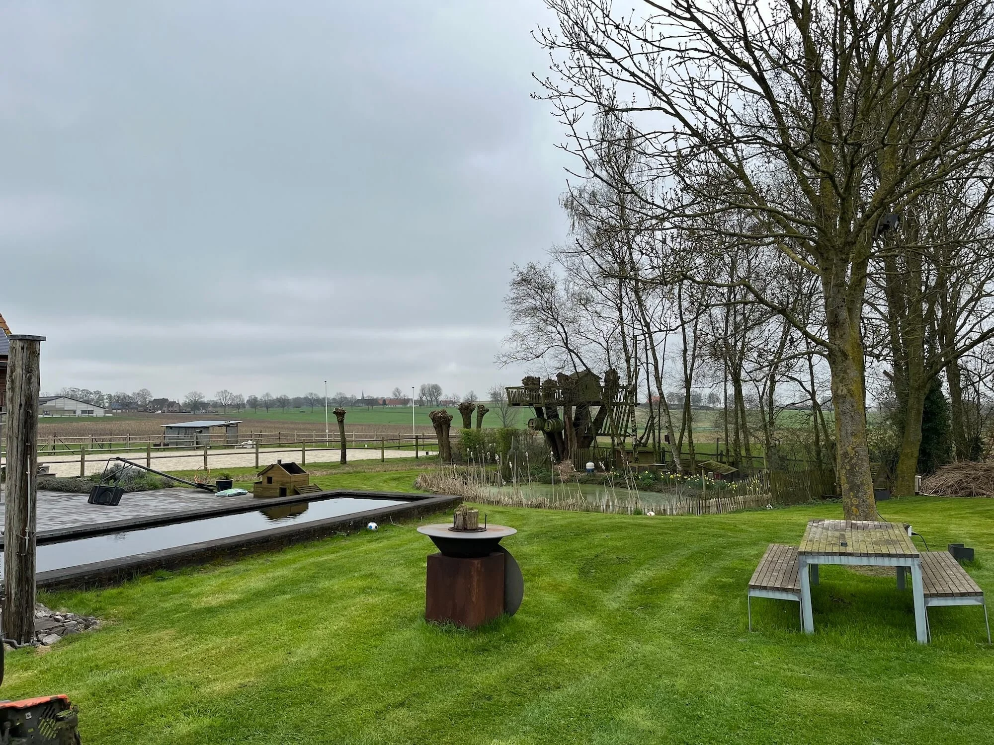 A backyard with a above-ground swimming pool, a small table, trees, a large wooden play structure in the distance, and open fields under a cloudy sky.