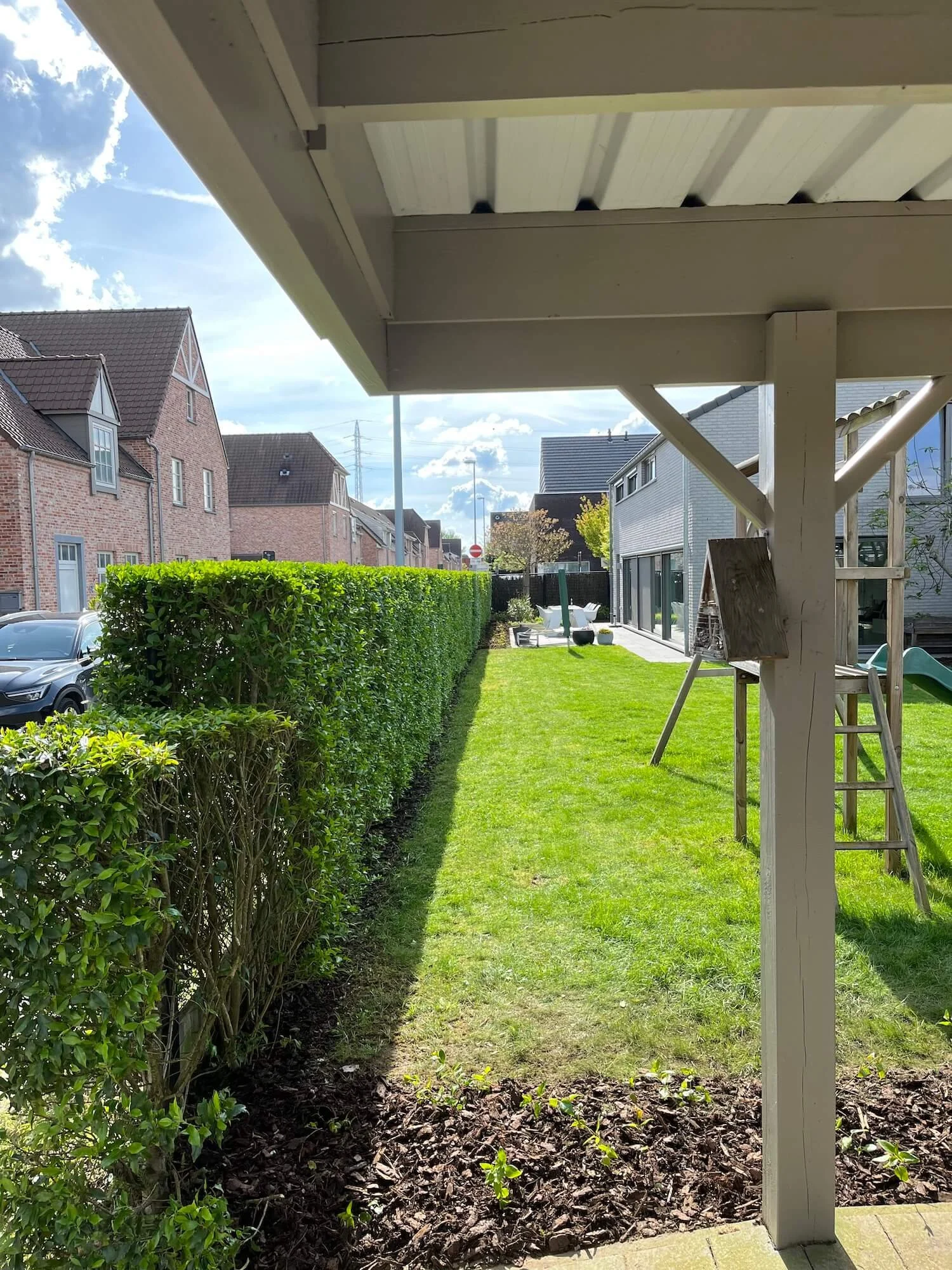 View from beneath a porch roof showing a backyard with a well-kept lawn, a tall hedge, outdoor furniture, a slide, and neighboring houses under a partly cloudy sky.