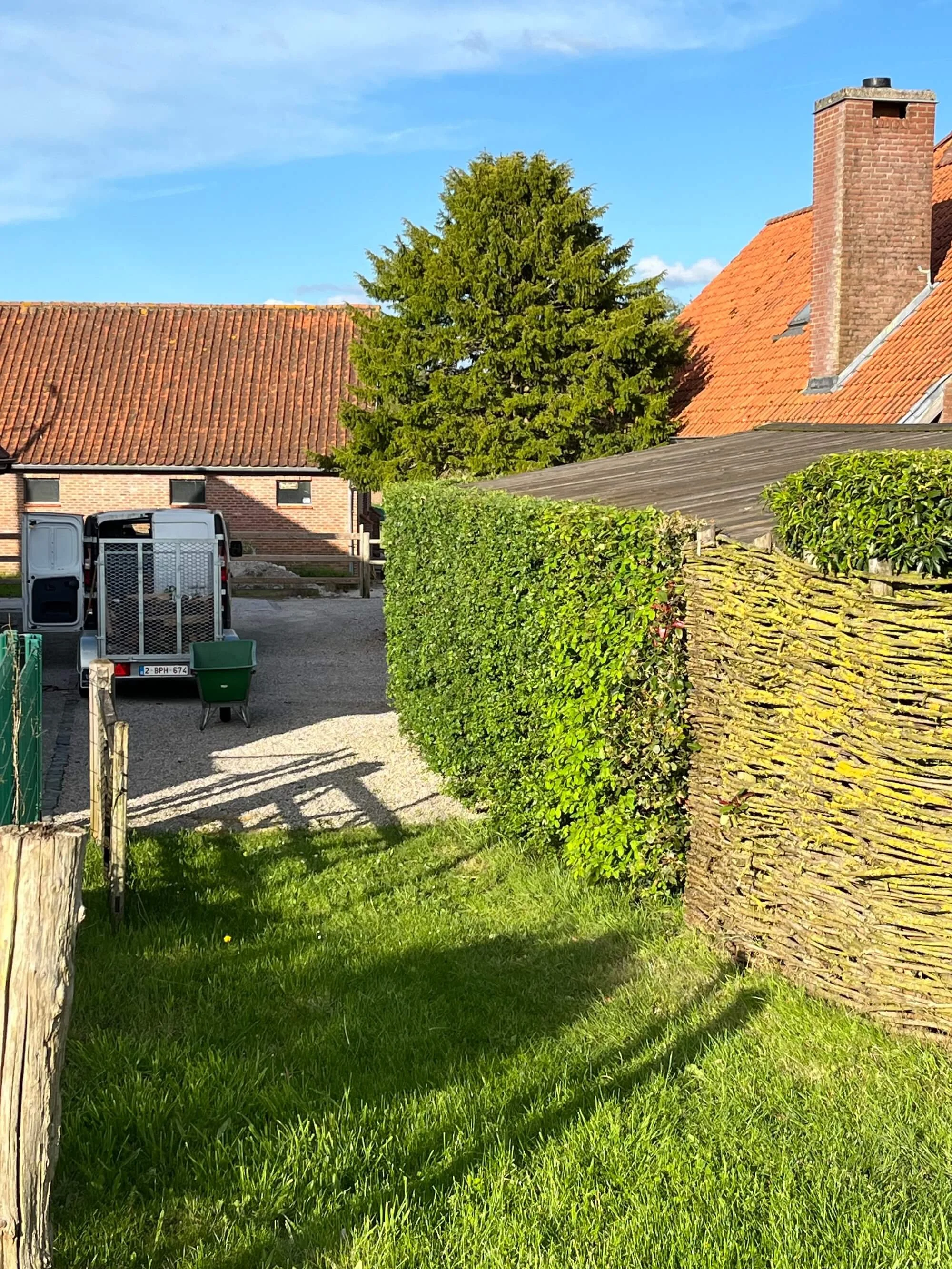 A gravel driveway with a white van parked, a green bin, surrounded by green bushes, a wooden and woven fence, a grassy area in the foreground, and red-roofed brick houses under a blue sky.