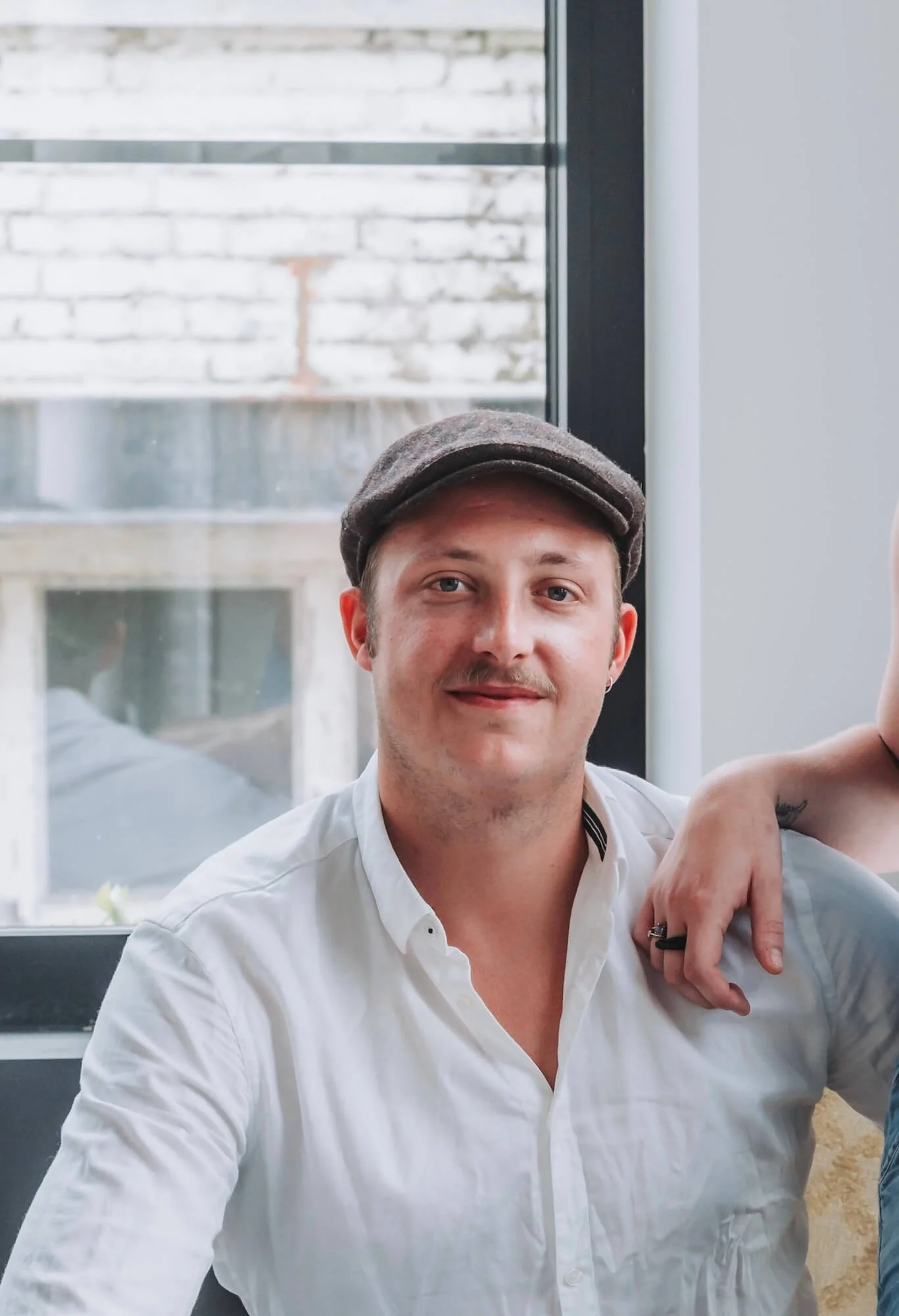 A man with light skin, wearing a white shirt and a brown cap, smiling, sitting indoors near a window.