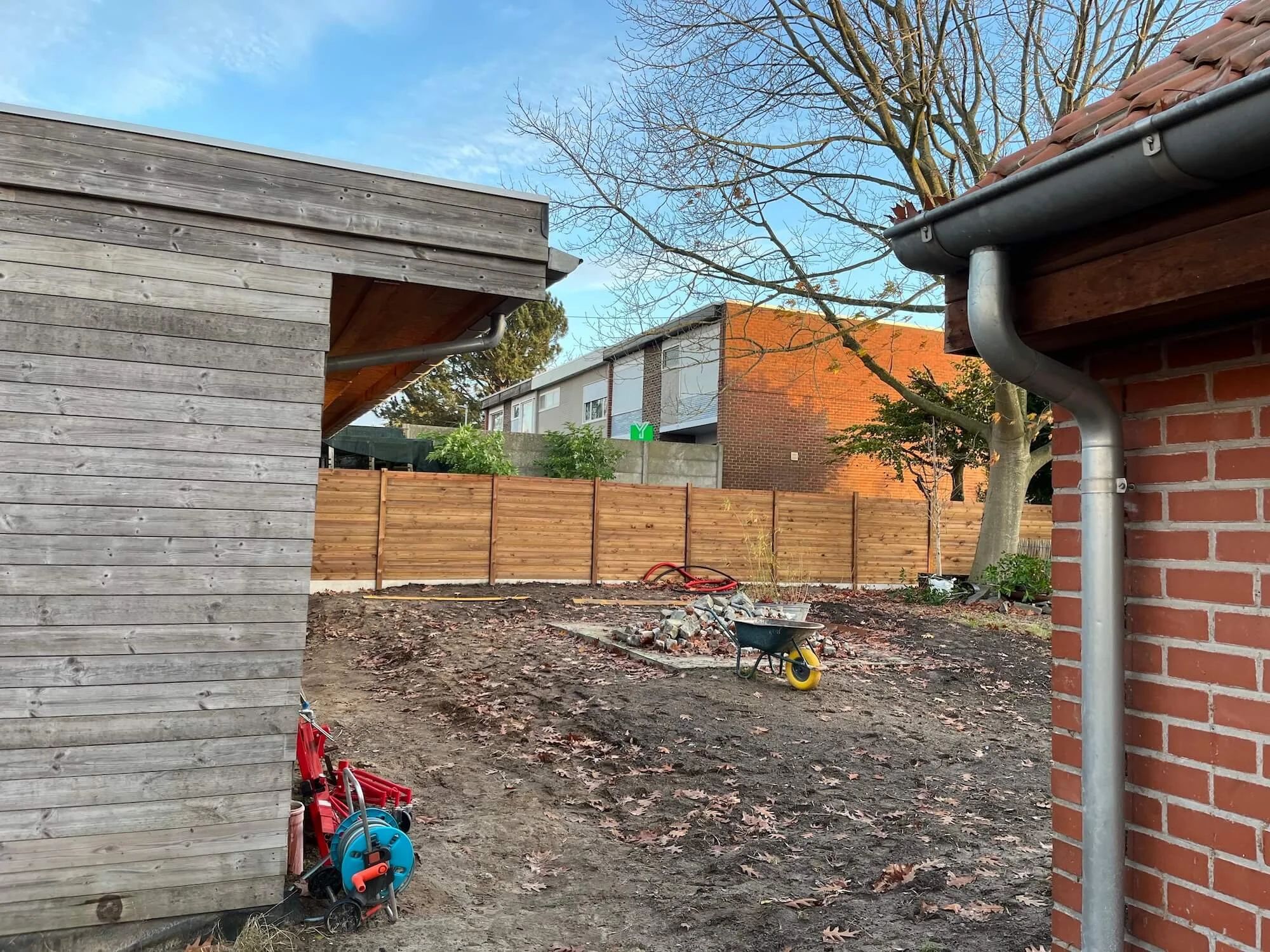 View of a backyard with a dirt ground, a wooden fence, a leafless tree, a wheelbarrow, and a small pile of bricks, with neighboring houses visible in the background and a clear sky overhead.