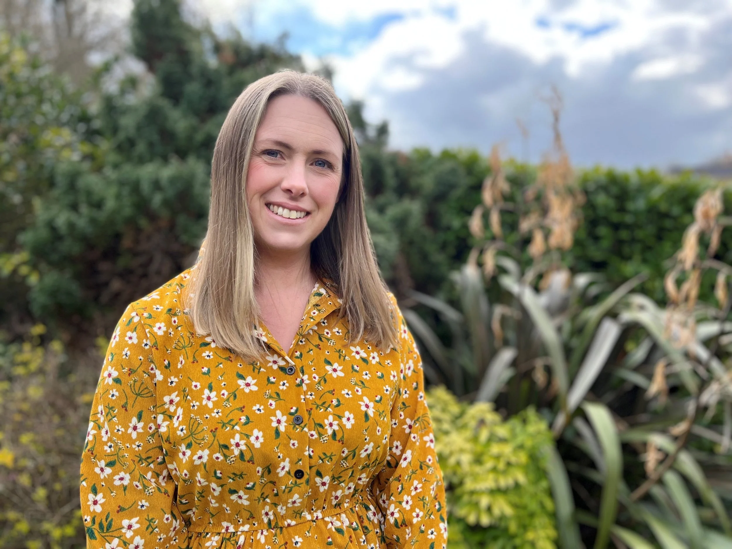 A woman with shoulder-length blonde hair, smiling, wearing a yellow floral dress, standing outdoors in a garden with green bushes and plants, under a partly cloudy sky.