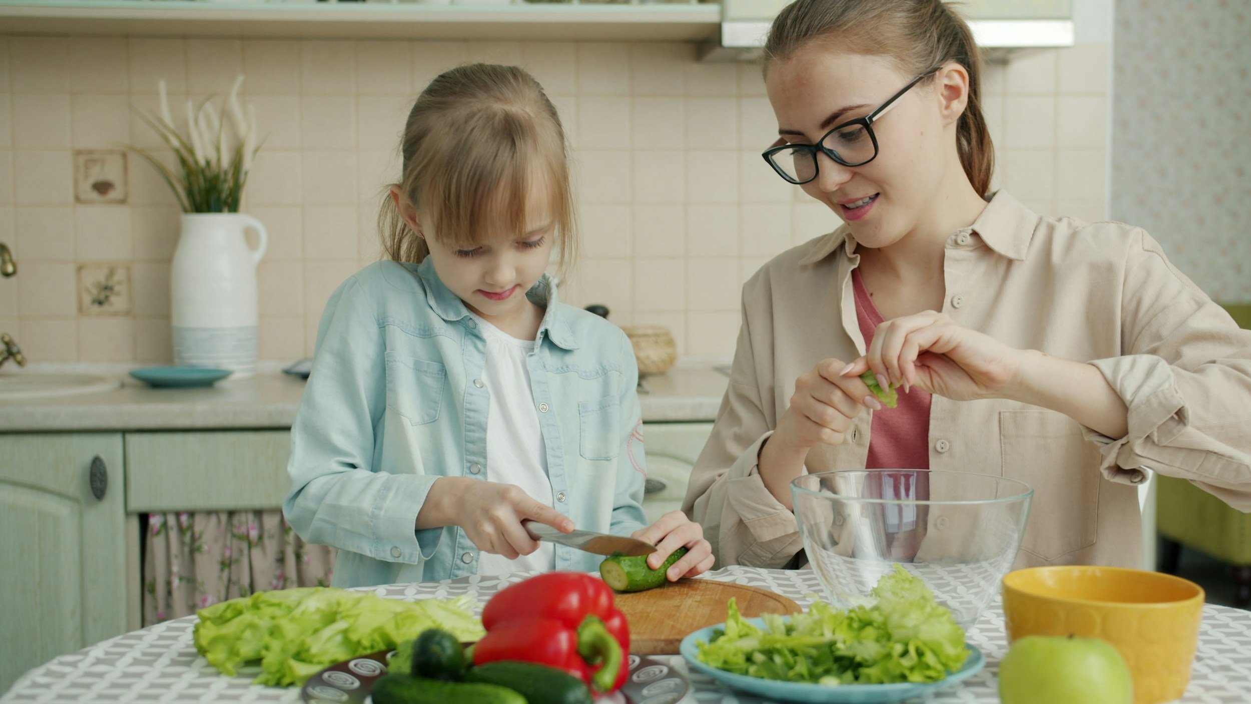 A woman and a young girl prepare a salad in a kitchen with fresh vegetables on the table, including cucumbers, lettuce, red bell peppers, and an apple.