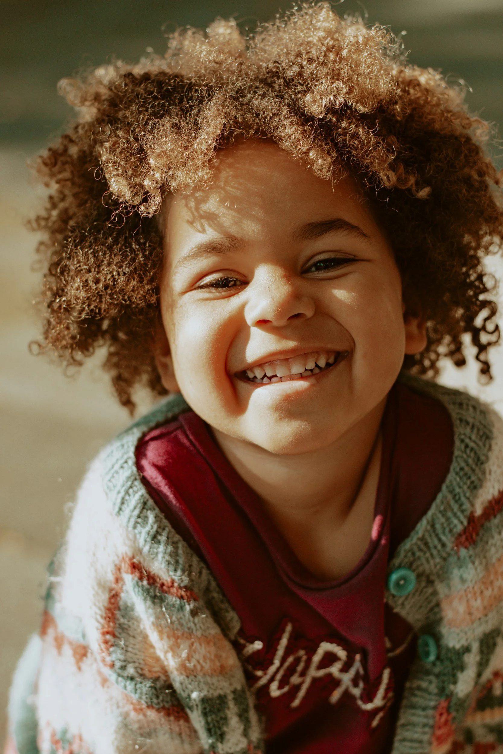 Close-up of a smiling young girl with curly brown hair, wearing a multicolored sweater and maroon shirt, outdoors on a sunny day.