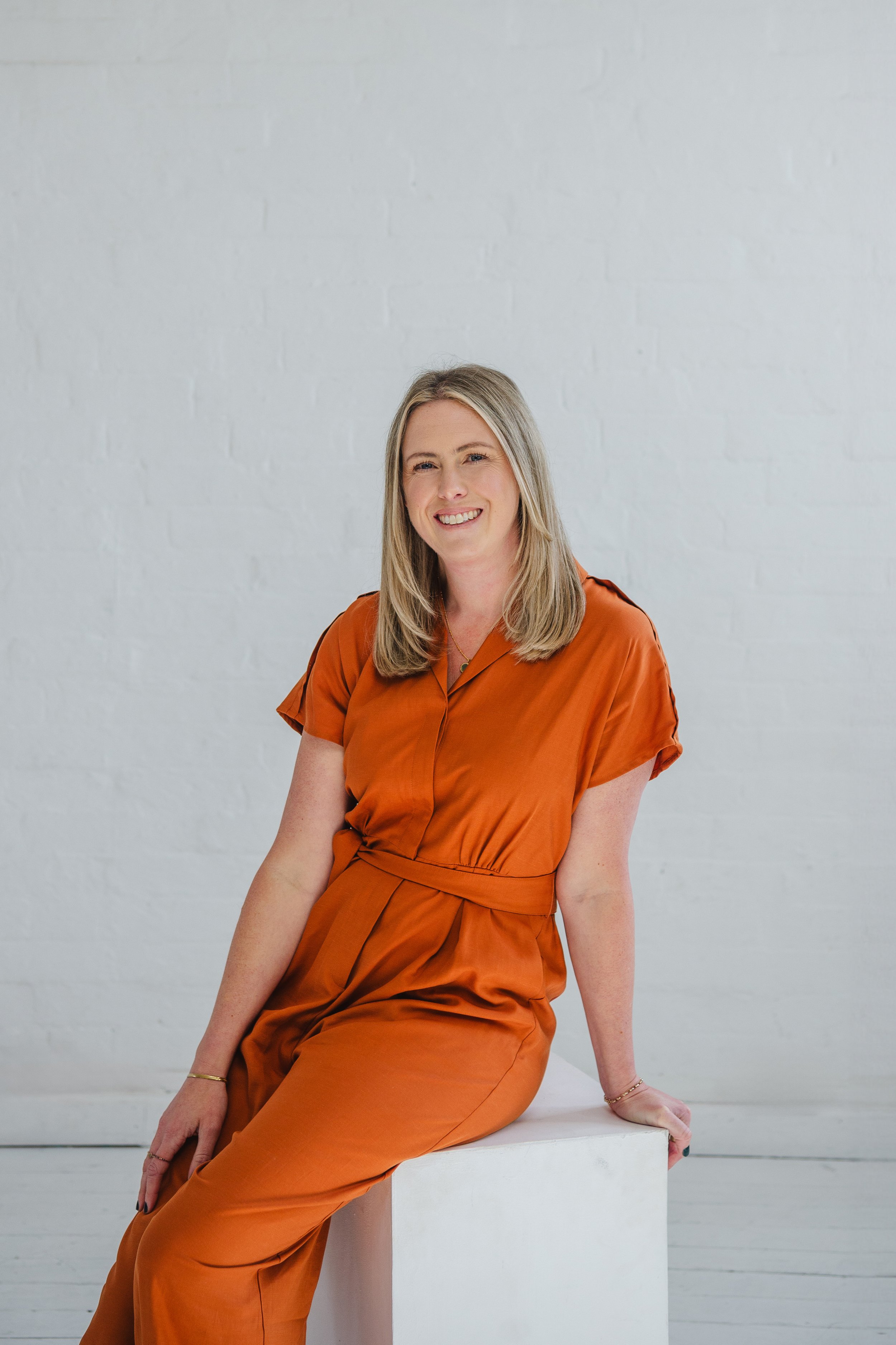 A smiling woman with blonde hair, wearing an orange dress, sitting on a white block against a white brick wall.