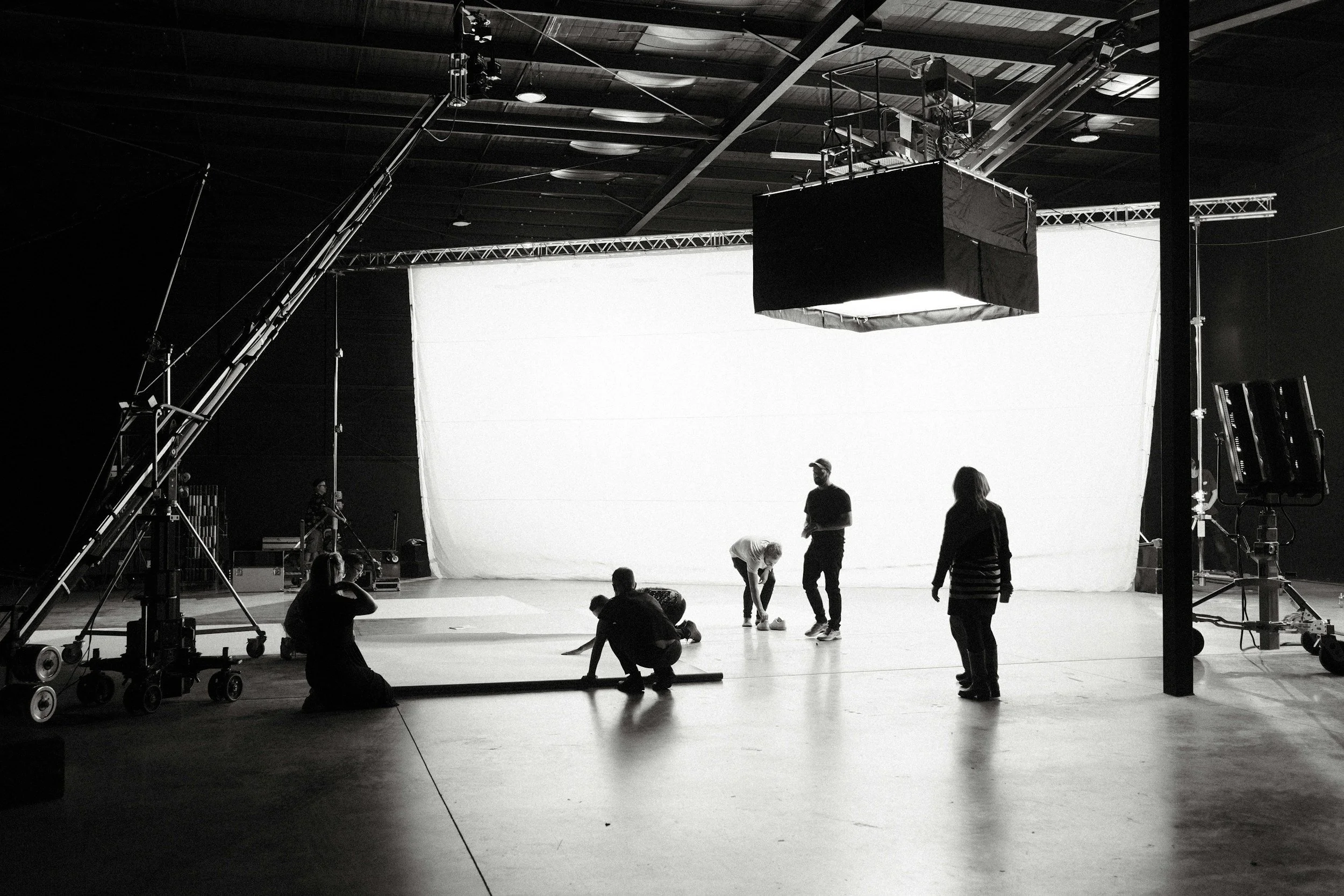 Film crew setting up on a black-and-white studio stage with a large white backdrop, including crew members handling equipment and preparing the scene.