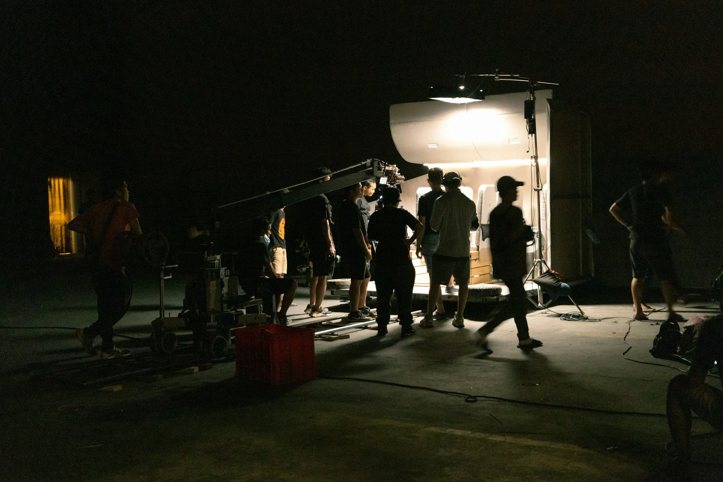 Film or television production crew working on a set at night, with lighting and camera equipment surrounding a white structure that resembles a small building or set piece.