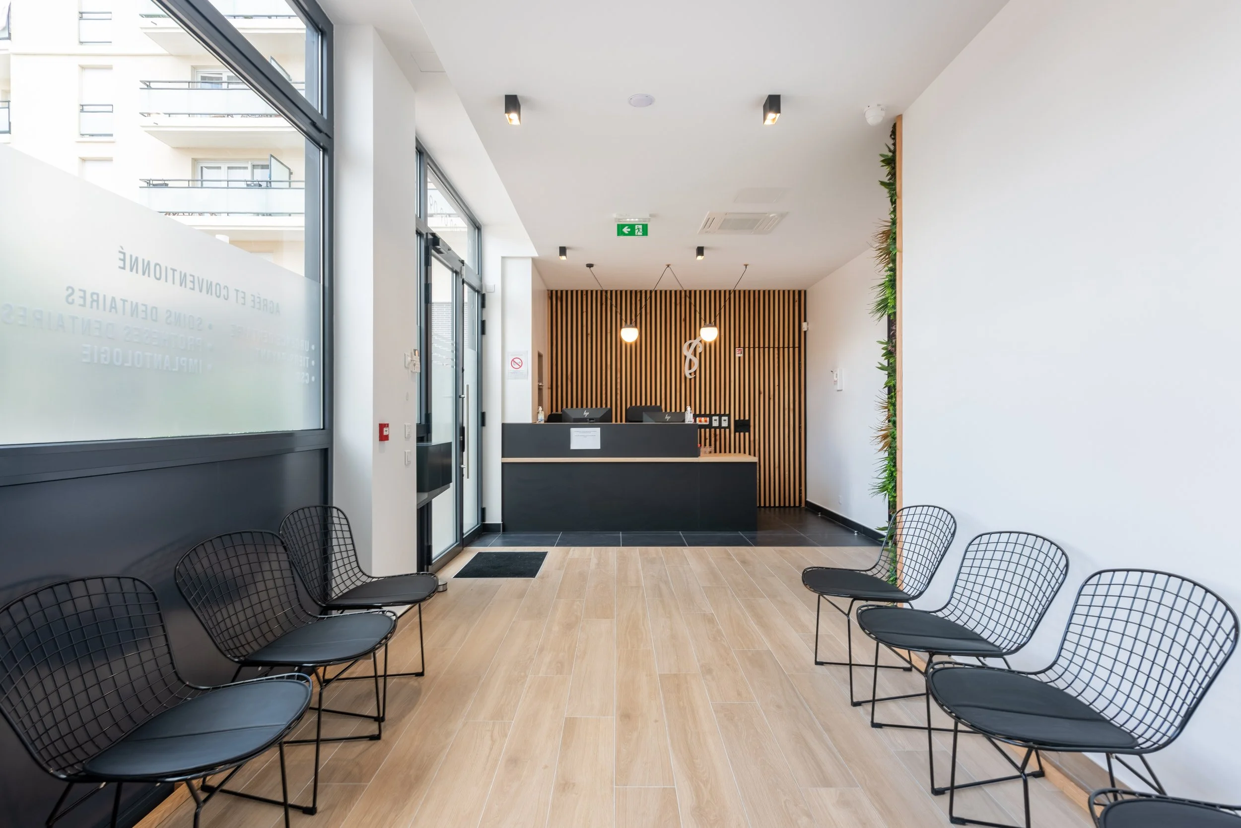 Salle d'attente moderne avec des chaises en métal noir, un comptoir de réception noir, un mur en bois vertical, et une décoration verte sur le côté droit.