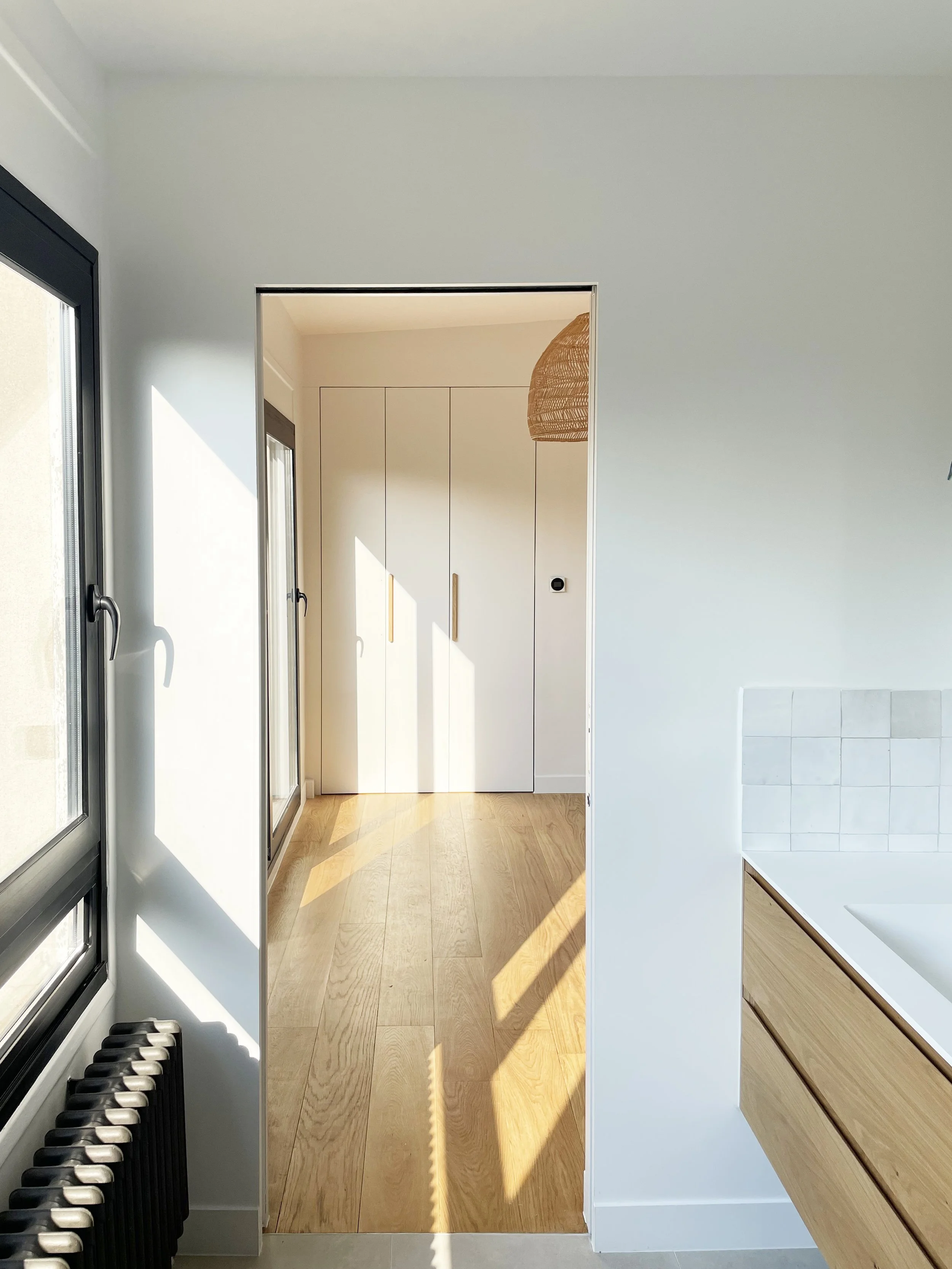 Intérieur lumineux d'une pièce moderne avec fenêtres, un radiateur noir, un meuble en bois clair avec des tiroirs, et une porte vers un espace avec des placards blancs.