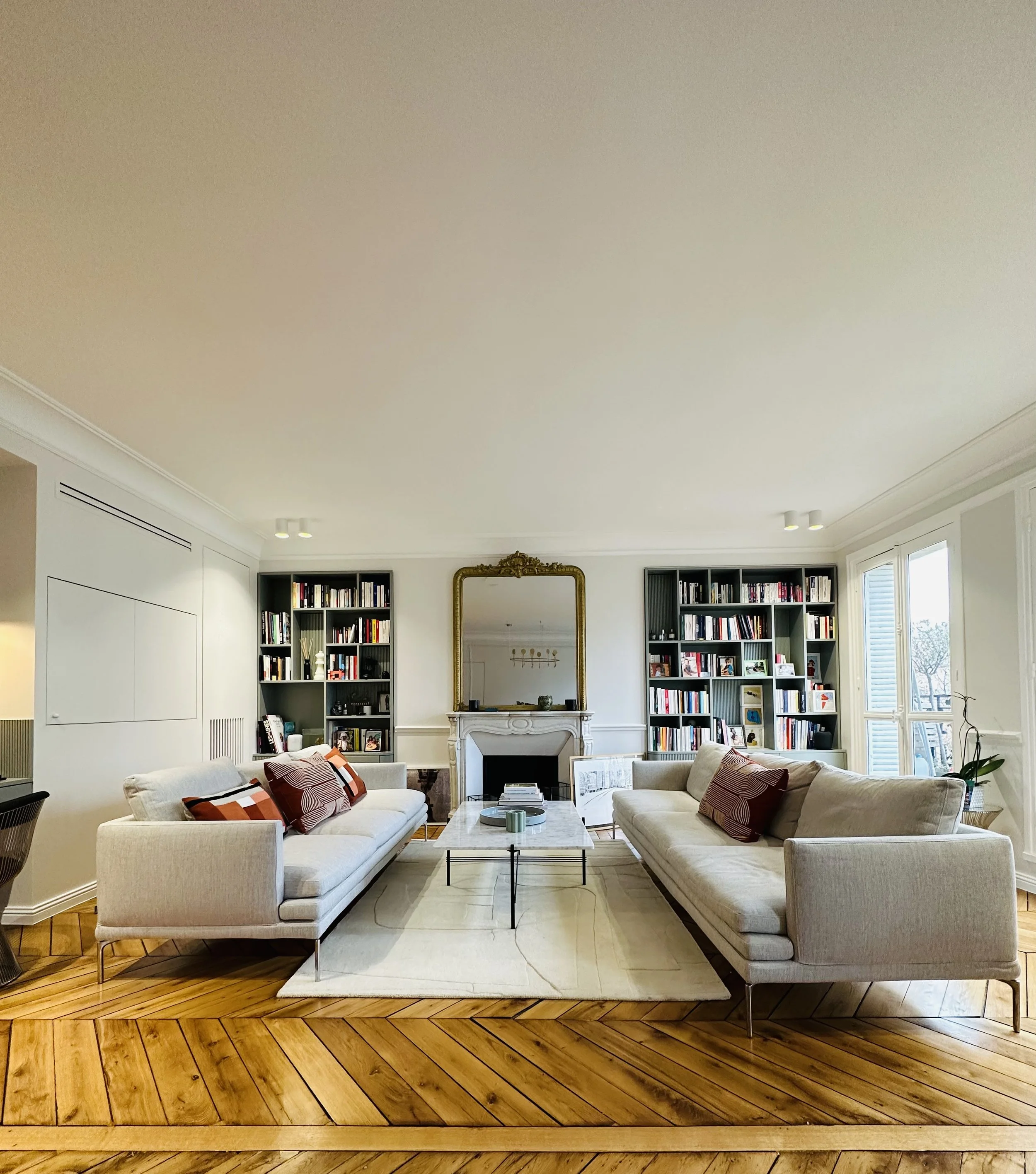 Salon élégant avec deux sofas beiges, un tapis clair, une cheminée avec un miroir doré au-dessus, deux étagères remplies de livres, et une grande fenêtre laissant entrer la lumière.