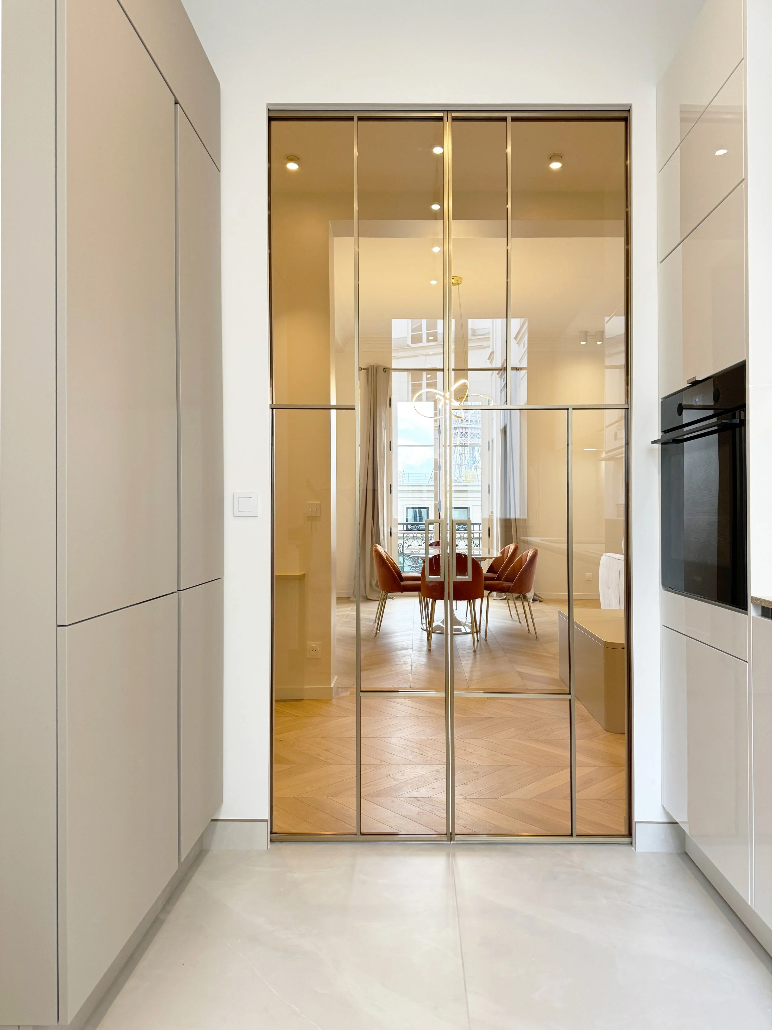 Une porte en verre miroir avec cadre en métal doré, séparant une cuisine moderne d'une salle à manger lumineuse avec fenêtre et chandelier.