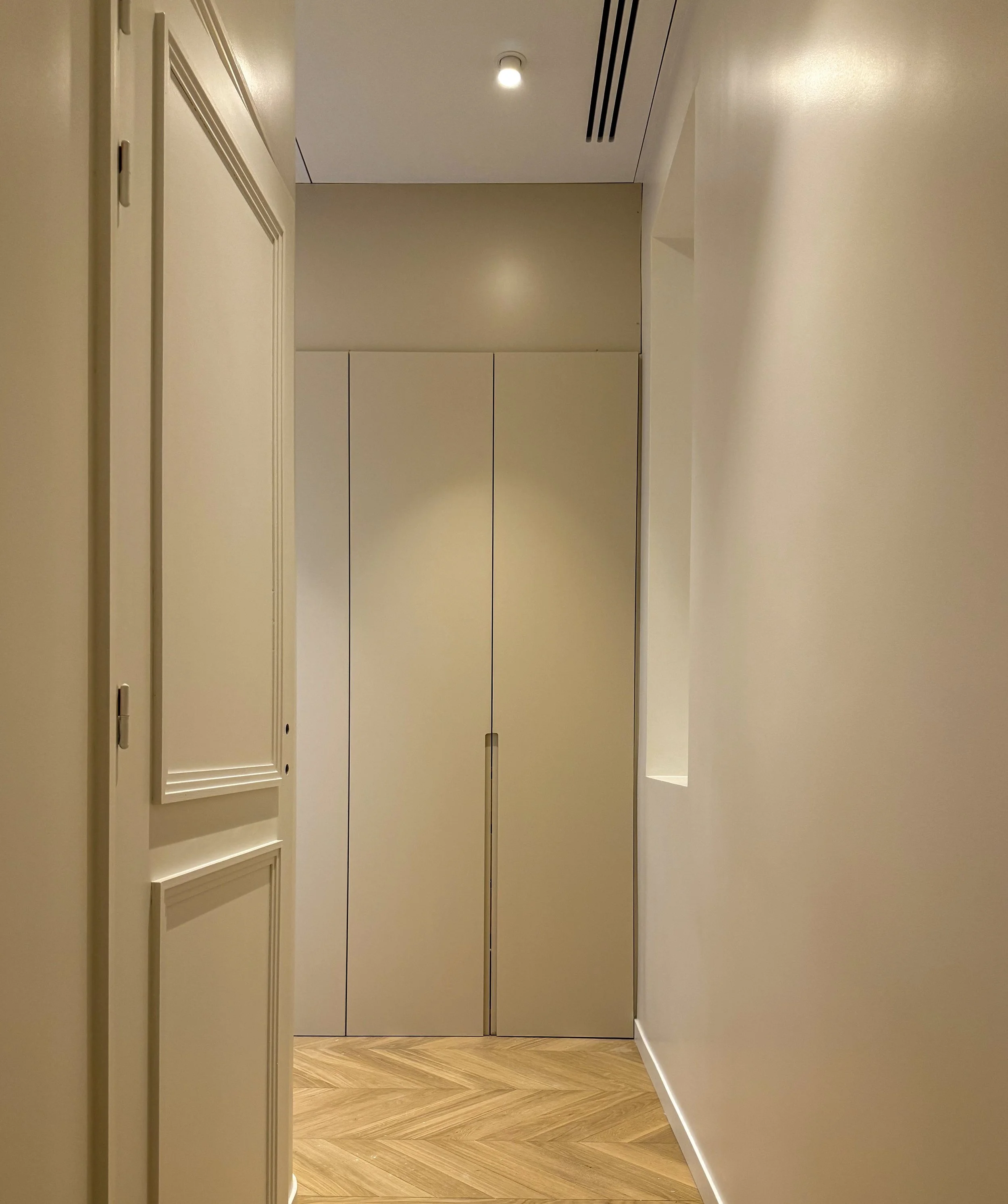Un couloir intérieur avec un sol en parquet, un mur beige à droite, un mur beige clair à gauche avec une porte en bois, et une armoire ou un placard beige au fond, avec un plafond blanc, un éclairage intégré et une ventilation.
