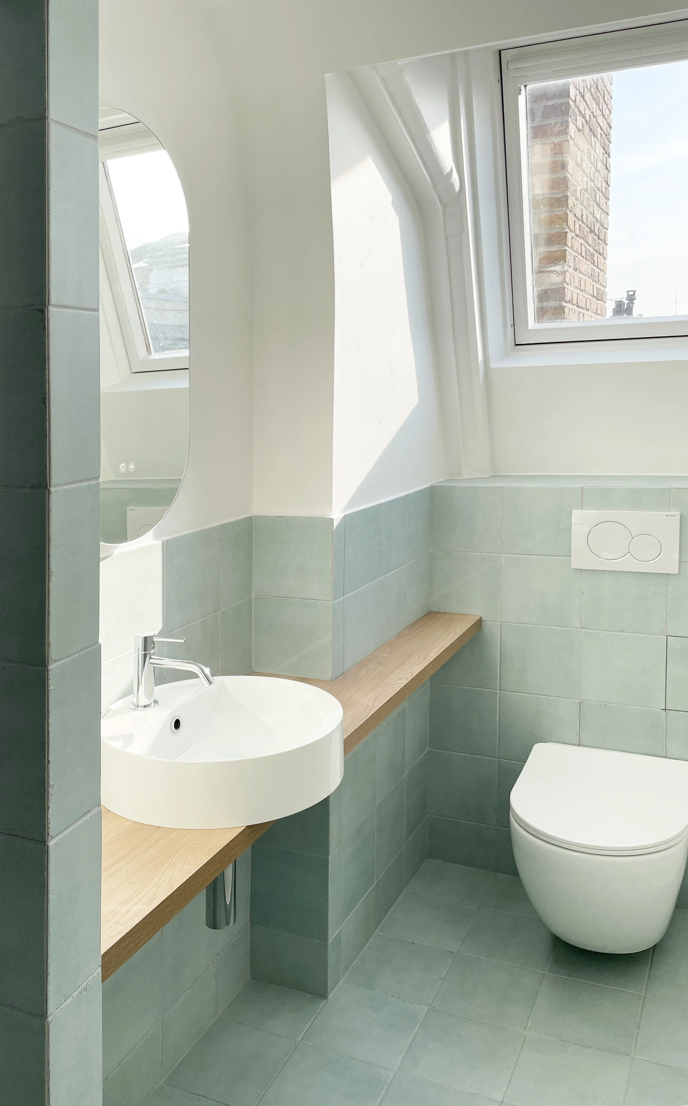 Une salle de bain moderne avec un lavabo rond blanc, un miroir ovale, un WC suspendu, un mur en carreaux vert pastel, et une fenêtre laissant entrer la lumière naturelle.