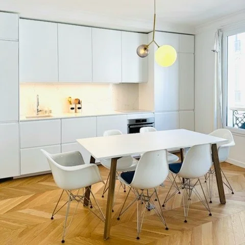 Cuisine moderne avec une table blanche et six chaises blanches autour, armoires blanches, parquet en bois, grande fenêtre avec rideaux, luminaire suspendu.