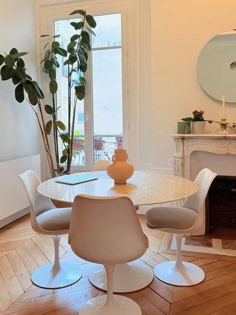 Salle à manger avec une table ronde en marbre, quatre chaises blanches, un grand plant à gauche, une fenêtre derrière, un rebord de cheminée décoré avec des fleurs et une chandelle, et un miroir rond sur le mur.