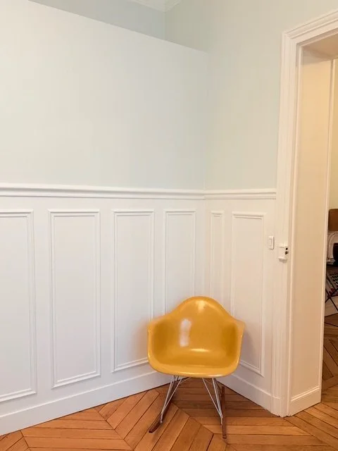 Une chaise jaune en plastique dans un coin d'une pièce avec un mur blanc et un panneau de bois au sol.