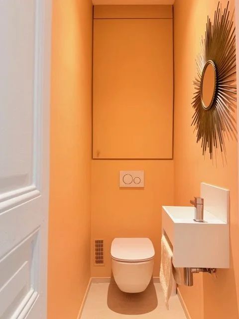 Petite salle de bain aux murs orange, équipée d'un WC suspendu, d'un petit lavabo et d'un miroir décoratif avec une étoile ou un soleil stylisé.