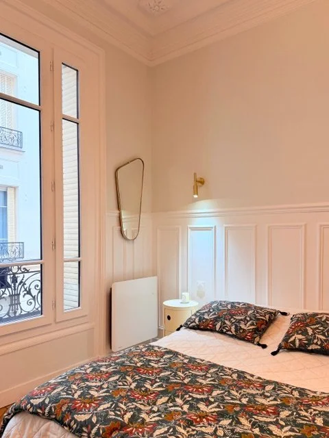 Chambre à coucher avec un lit, deux oreillers à motifs floraux, une petite table de chevet avec une lampe, une grande fenêtre, un miroir accolé au mur, et un décor intérieur élégant.