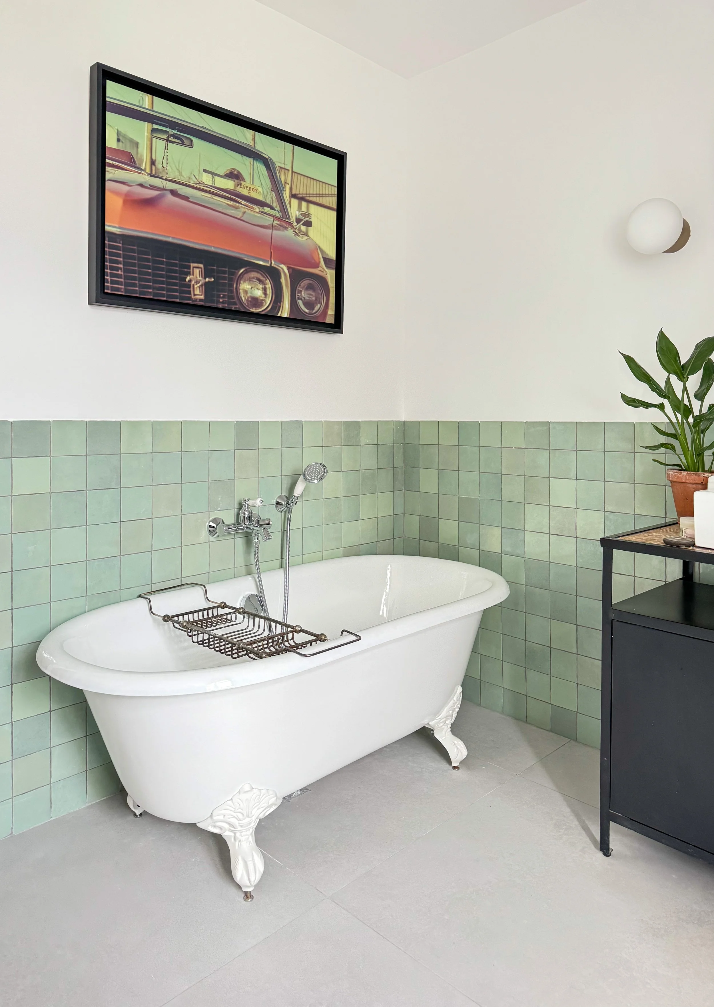 Une baignoire ancienne blanche avec pattes décoratives, située dans une salle de bain avec des carreaux verts au mur. Au-dessus, une affiche de voiture vintage et une plante verte dans un pot en terre cuite sur une petite étagère noire.