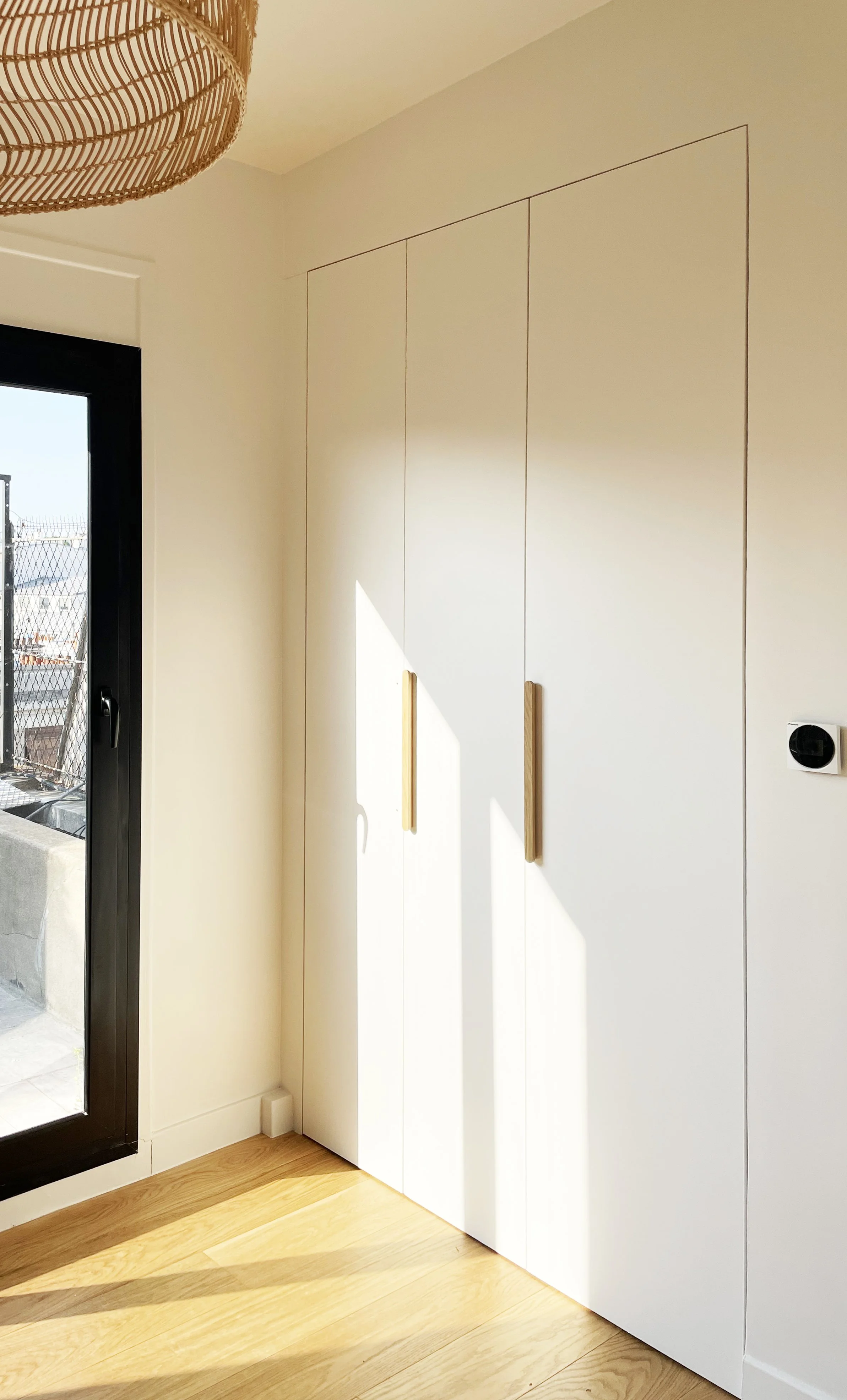 Une armoire blanche avec deux poignées en bois, à côté d'une porte-fenêtre donnant sur un balcon, avec un sol en bois et un plafond clair, et une lampe en rotin suspendue au plafond.