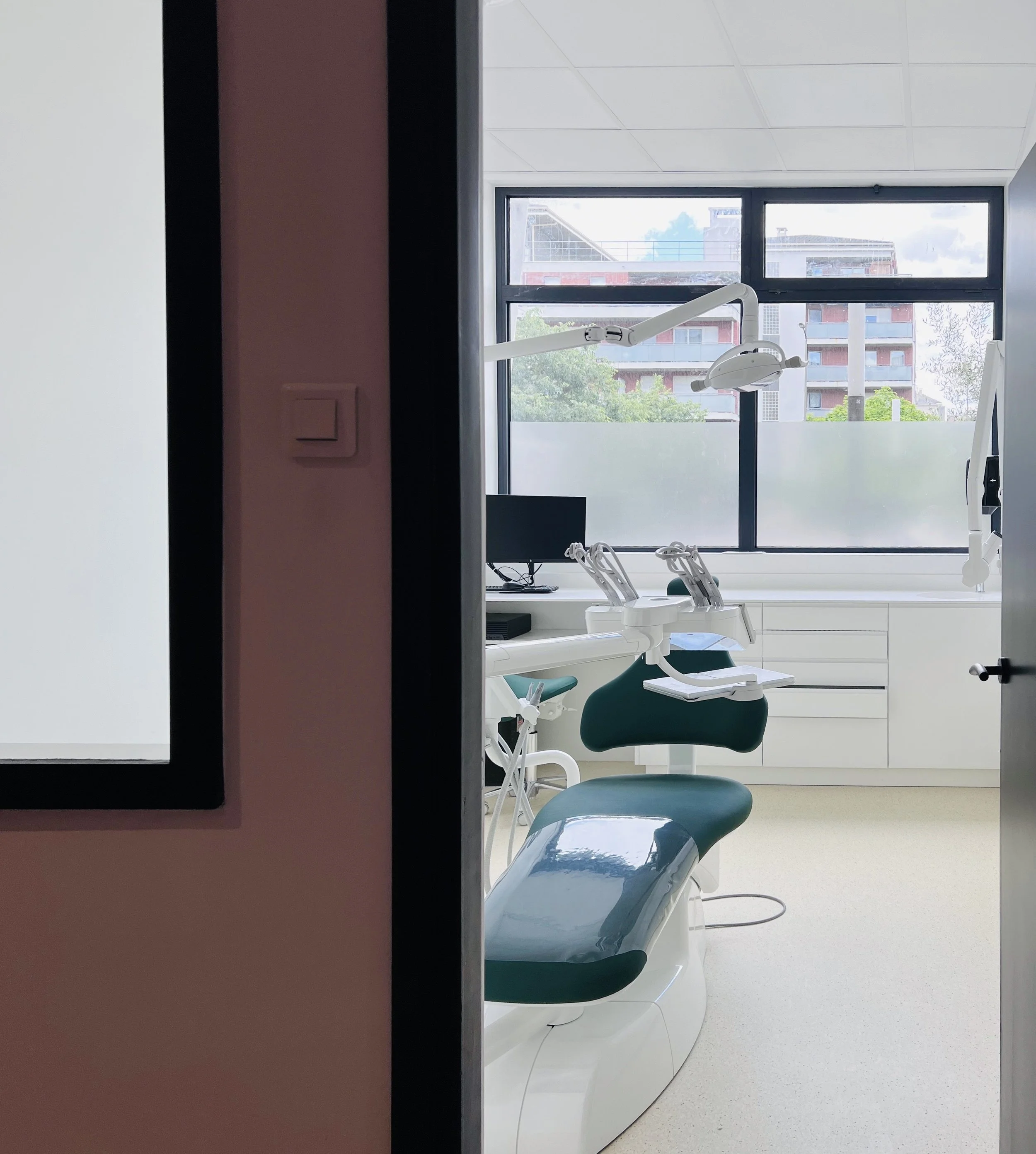 Une salle de soins dentaires avec un fauteuil de dentiste, un moniteur d'ordinateur, et une lumière d'examen, vue à travers une porte entrouverte