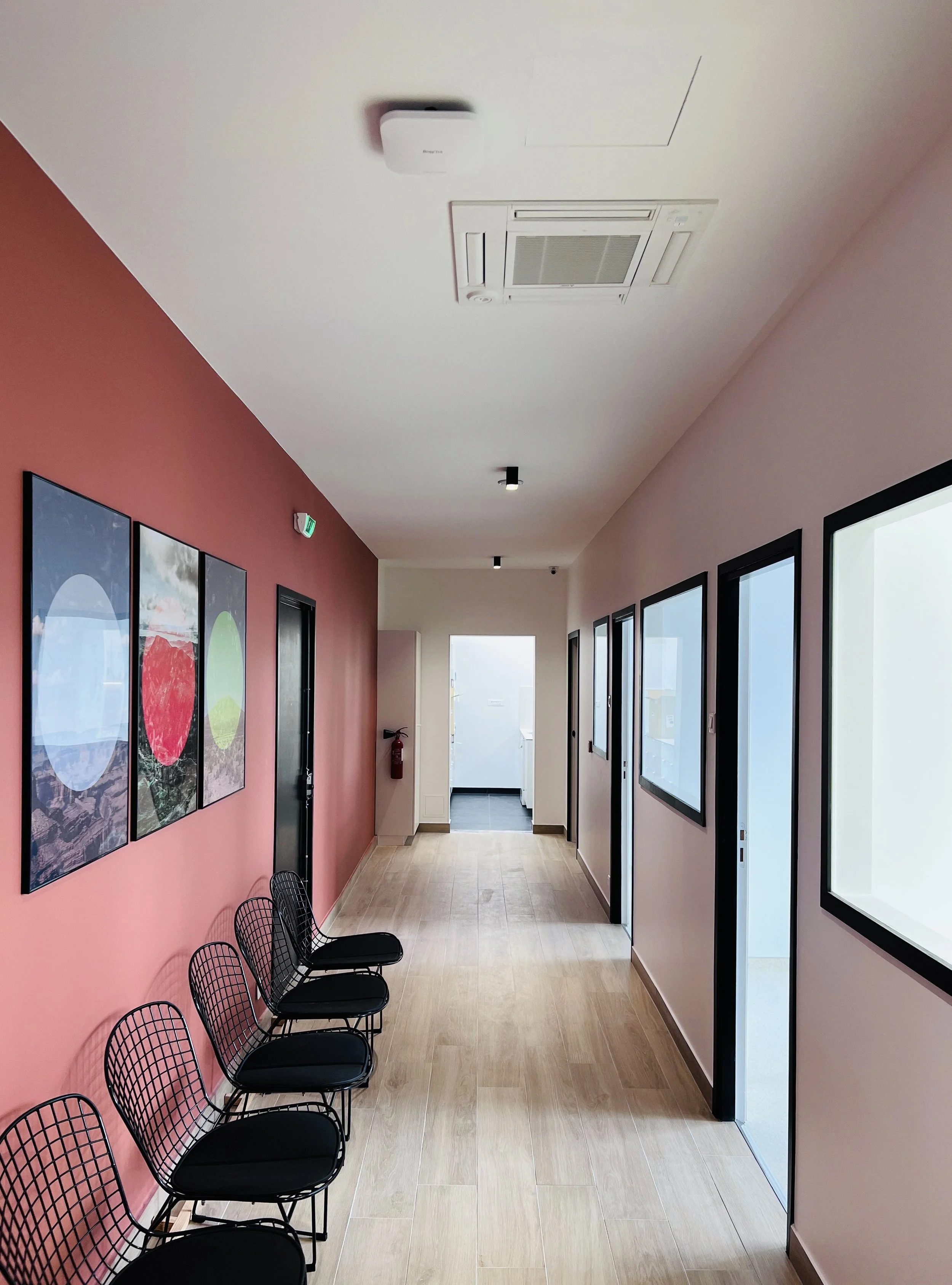 Couloir intérieur d'un bâtiment avec quatre chaises noires en métal, mur rose, trois œuvres d'art modernes, fenêtres et portes vitrées, sortie de secours avec éclairage, et porte de sortie visible au fond.