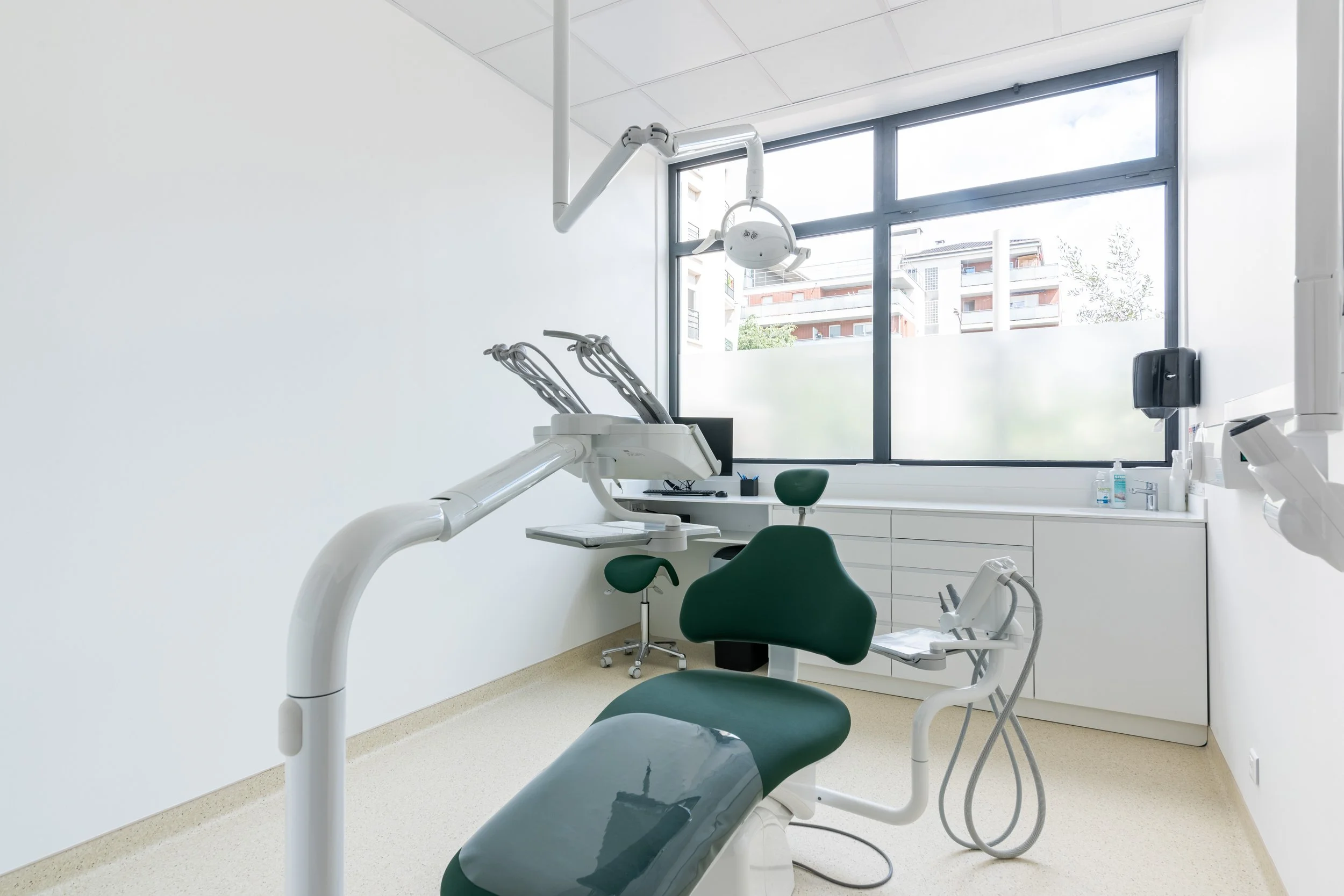 Salle d'examen dentaire avec une chaise de dentiste verte, appareils dentaires, grande fenêtre laissant entrer la lumière naturelle et mobilier blanc minimaliste.