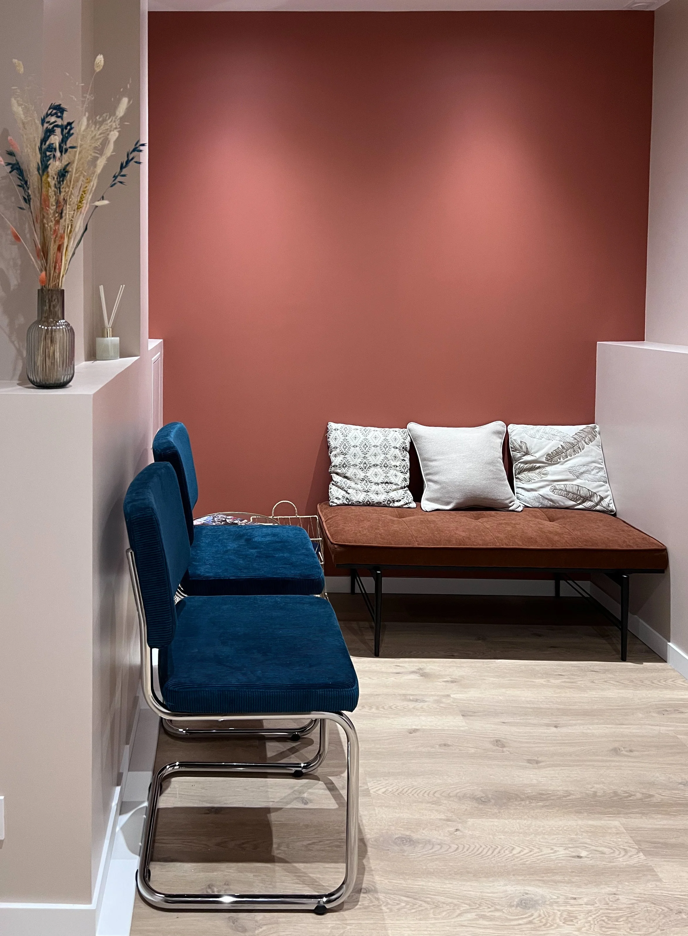 Un coin salon avec un canapé en arrière-plan avec trois coussins, un mur rose en accent, et trois chaises bleues en premier plan, avec un vase de fleurs séchées sur une étagère à gauche.