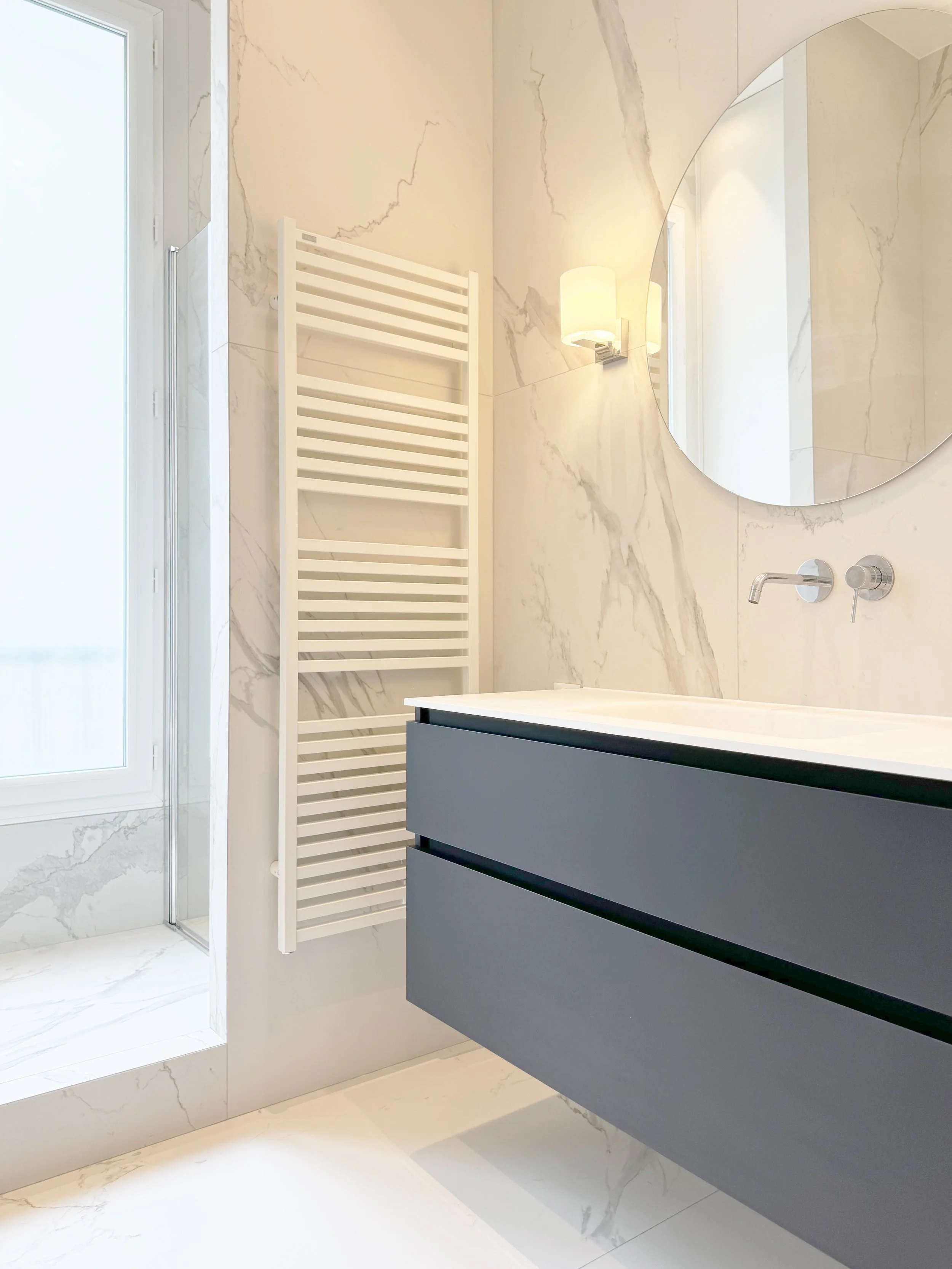 Salle de bain moderne avec un miroir rond, un meuble vasque gris foncé, un robinet mural, un miroir, un radiateur électrique et une fenêtre avec un carrelage en marbre blanc.