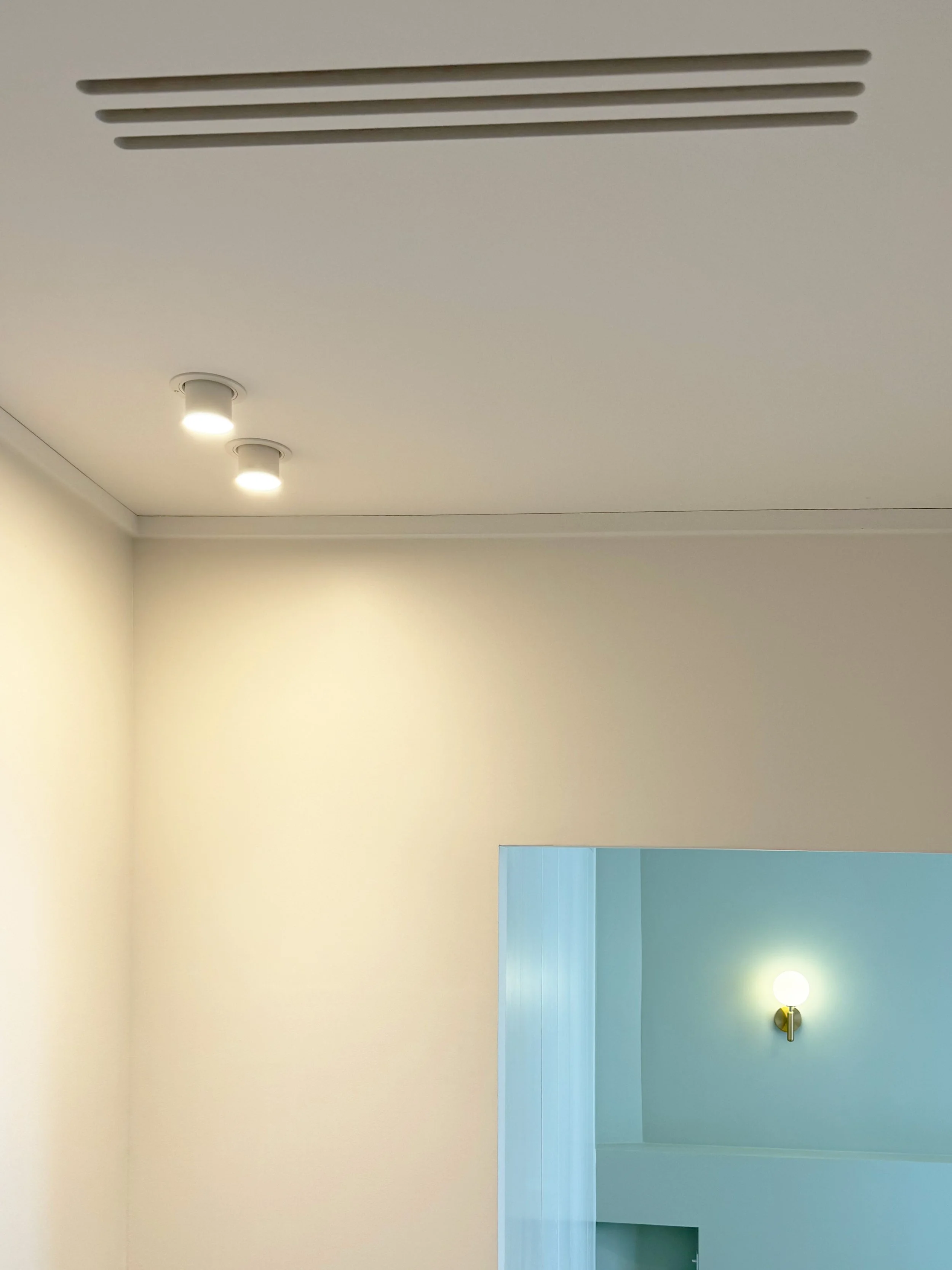 Plafond blanc avec deux spots de lumière encastrés et une grille d'aération. Mur blanc avec une ouverture menant à une pièce avec une lumière en forme de globe.