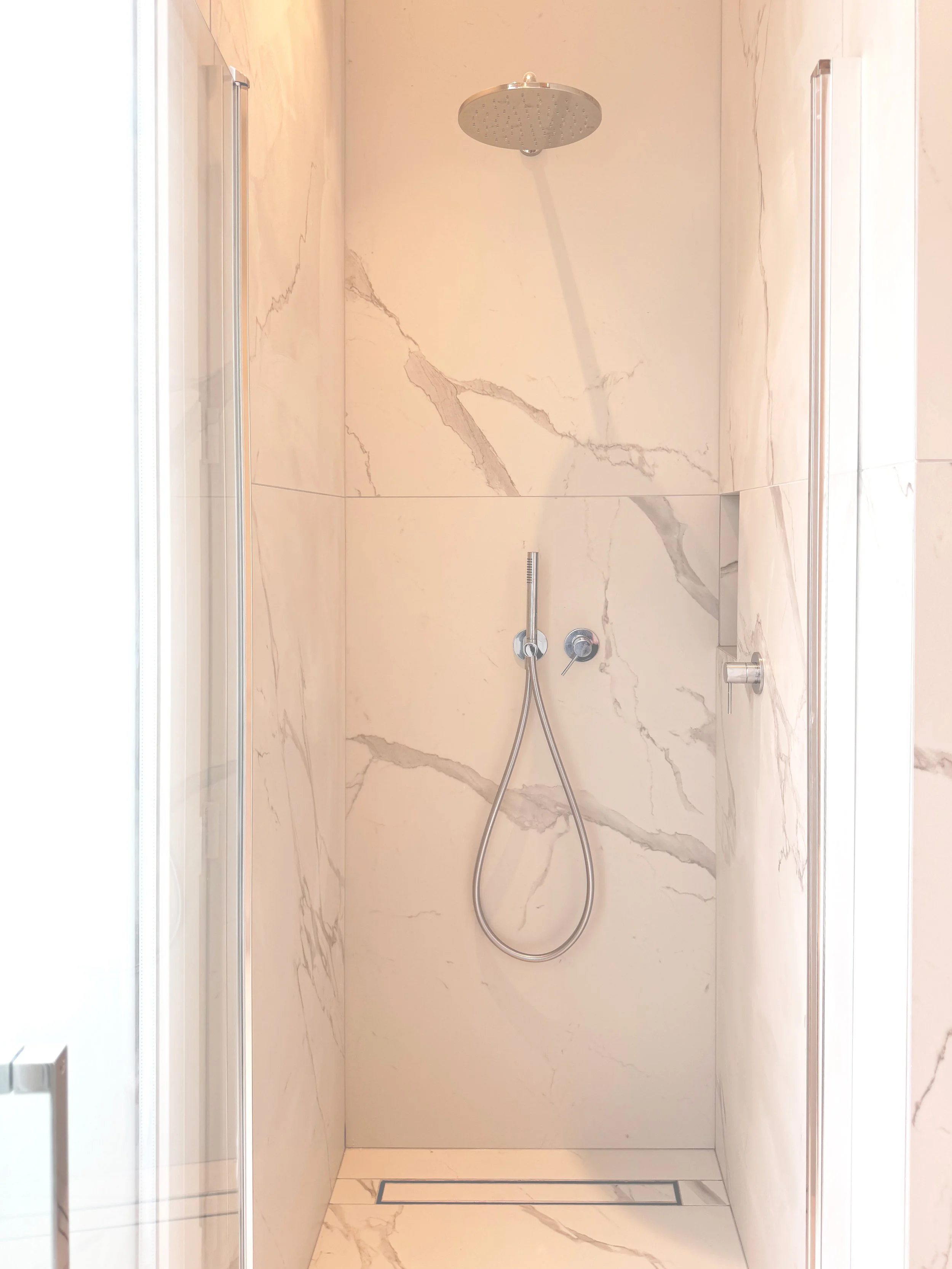 Douche à cabine avec un pommeau de douche ronde fixe au plafond, un distributeur de savon ou shampoing fixé à la muraille, parois en marbre blanc avec veines grises, conduit de drainage au sol.