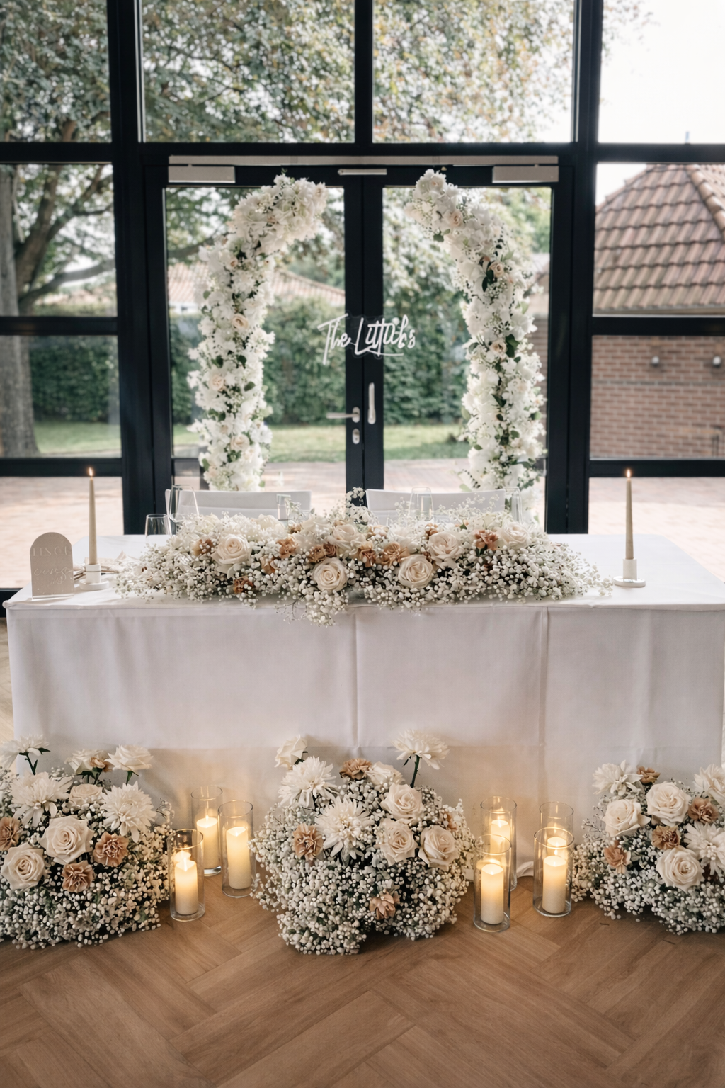 Elegant dekorierter Hochzeits- oder Eventtisch mit weißen und roséfarbenen Blumen, Kerzen in Glasgefäßen und floralem Fensterbogen.