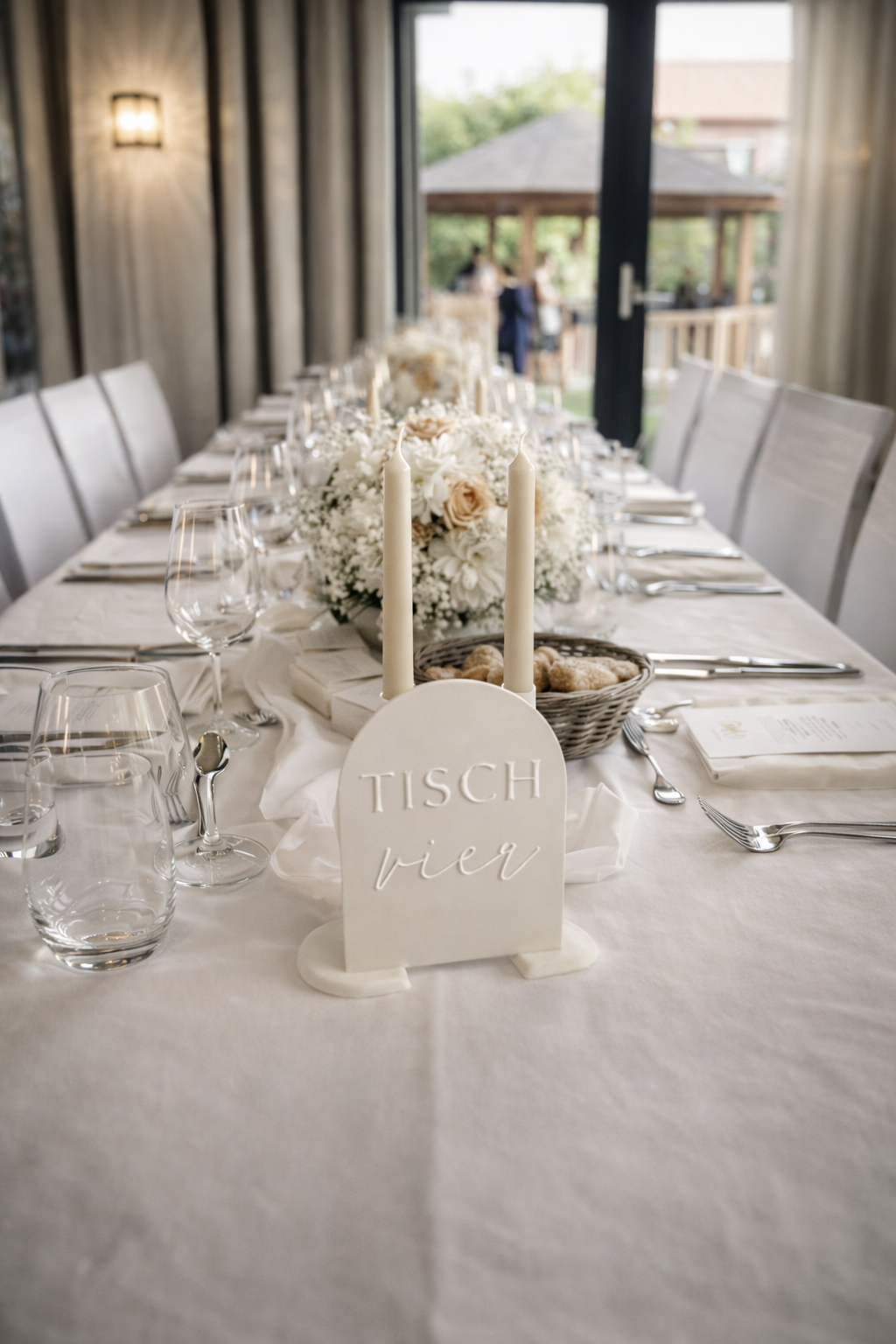 Festlich gedeckter Tisch mit weißer Hochzeitsdekoration, Kerzen, floralen Arrangements und Tischschild „Tisch vier“.