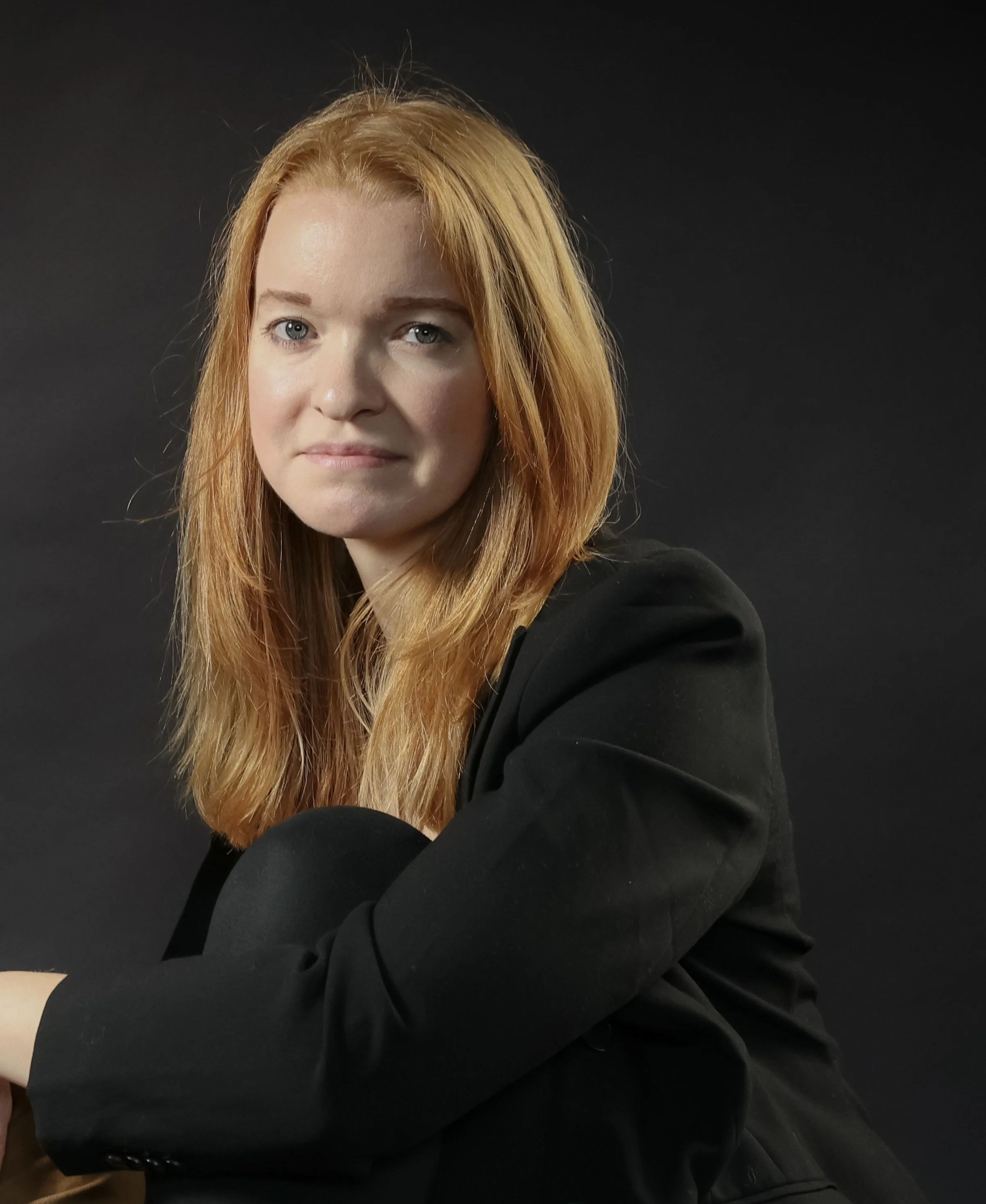 Porträt einer Frau mit schulterlangem roten Haar in schwarzem Blazer vor dunklem Hintergrund.