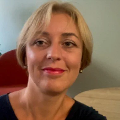 A woman with short blond hair smiling indoors, sitting in front of a light-colored wall and a round wooden table.